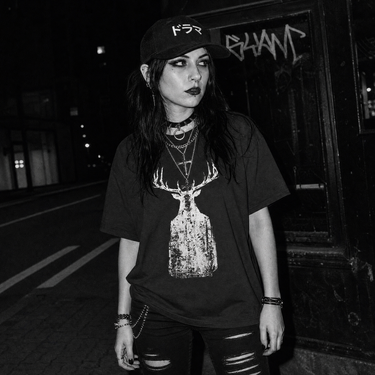 A woman wearing a black graphic t-shirt with a distressed deer on a ghostly human body print, a black cap, and ripped black jeans, standing on a dark street at night.