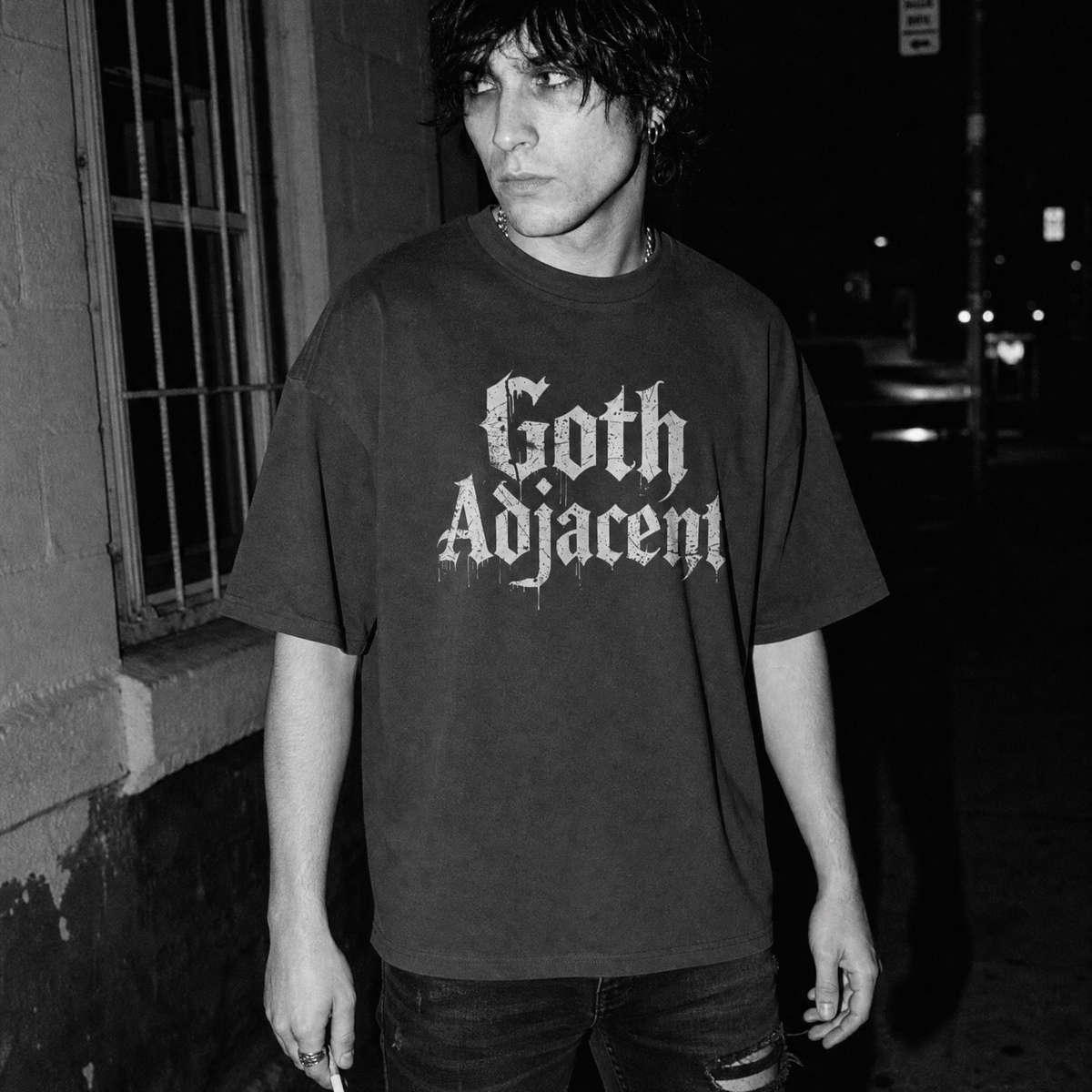 A man standing outdoors at night wearing a dark oversized T-shirt with the words 'Goth Adjacent' printed in a large white gothic-style font across the chest.