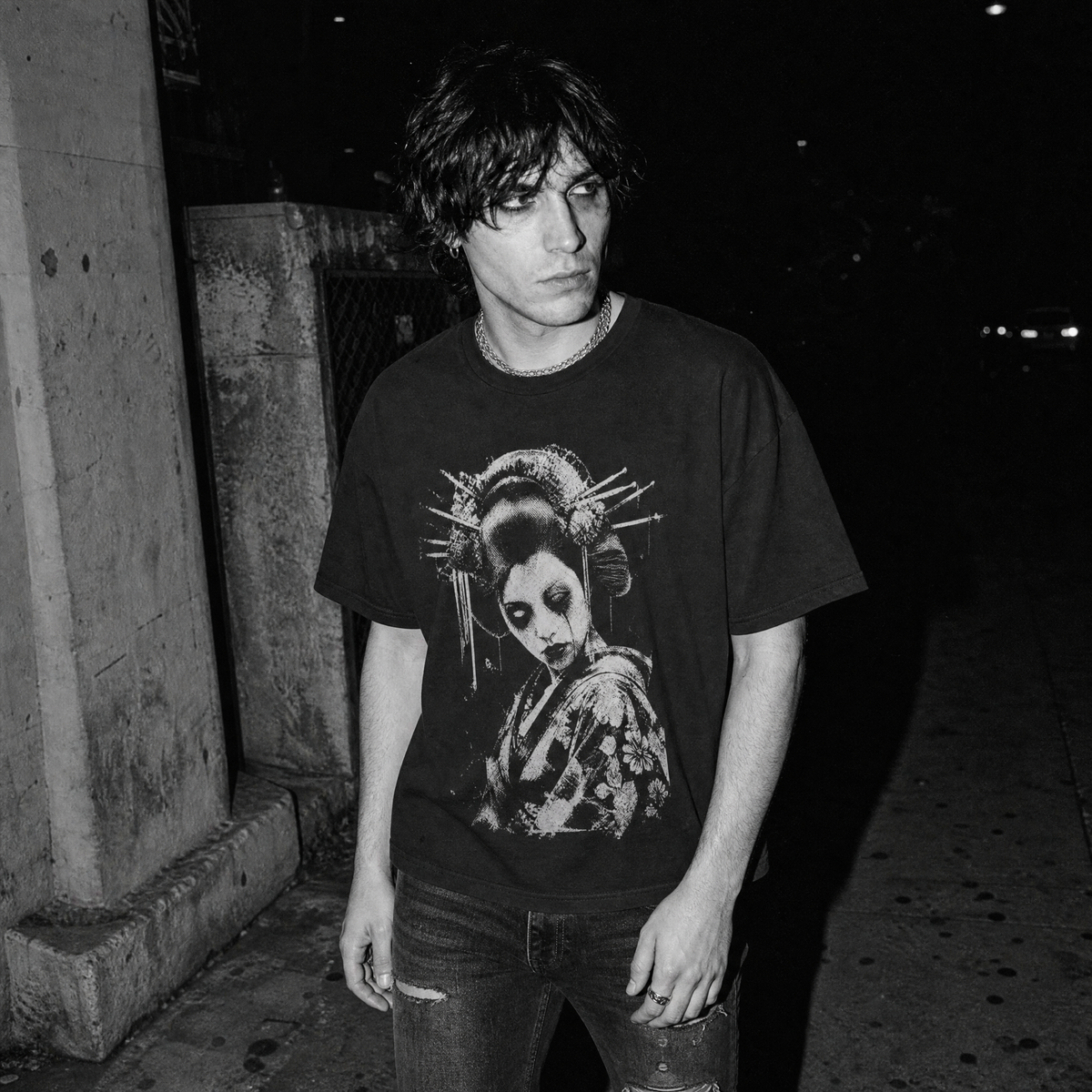 A man wearing a black graphic t-shirt with a high-contrast distressed print of a Geisha, photographed in black and white at night.