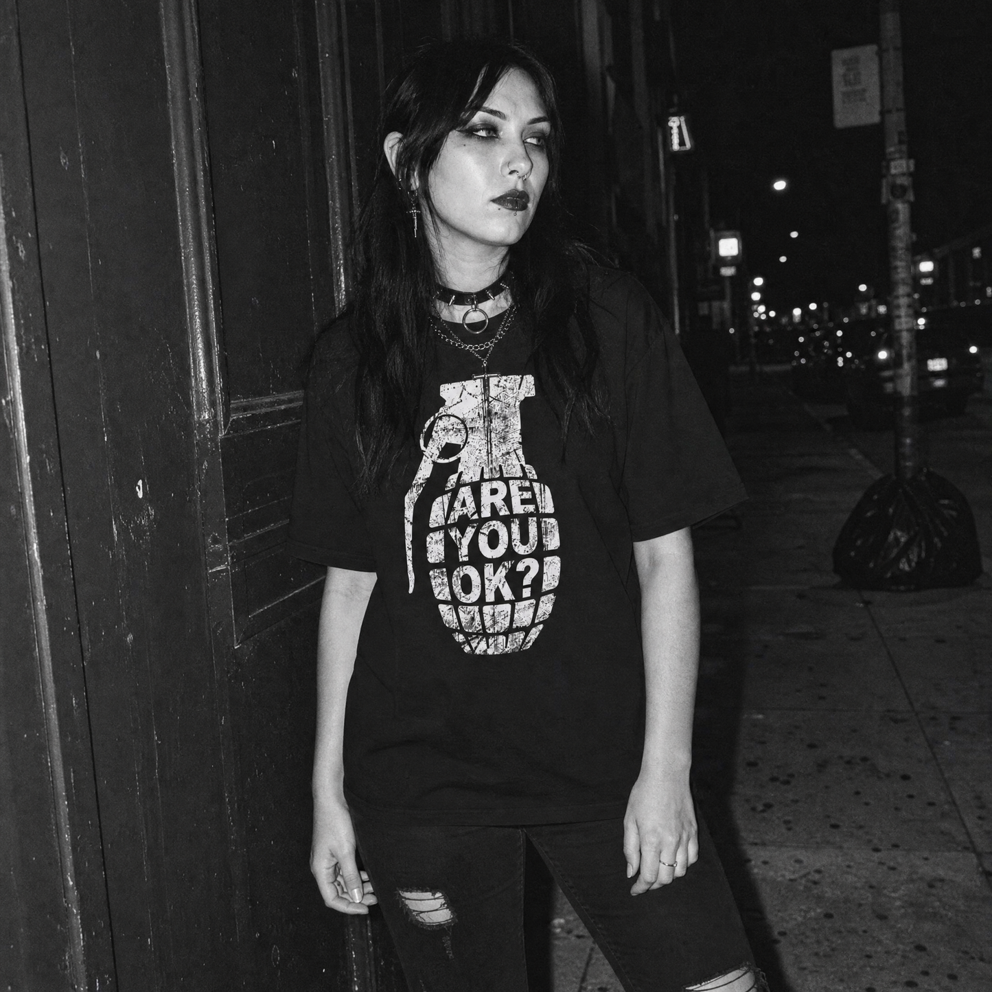 A woman wearing a black graphic t-shirt with a white grenade design and the text 'ARE YOU OK?' stands on a city sidewalk at night.
