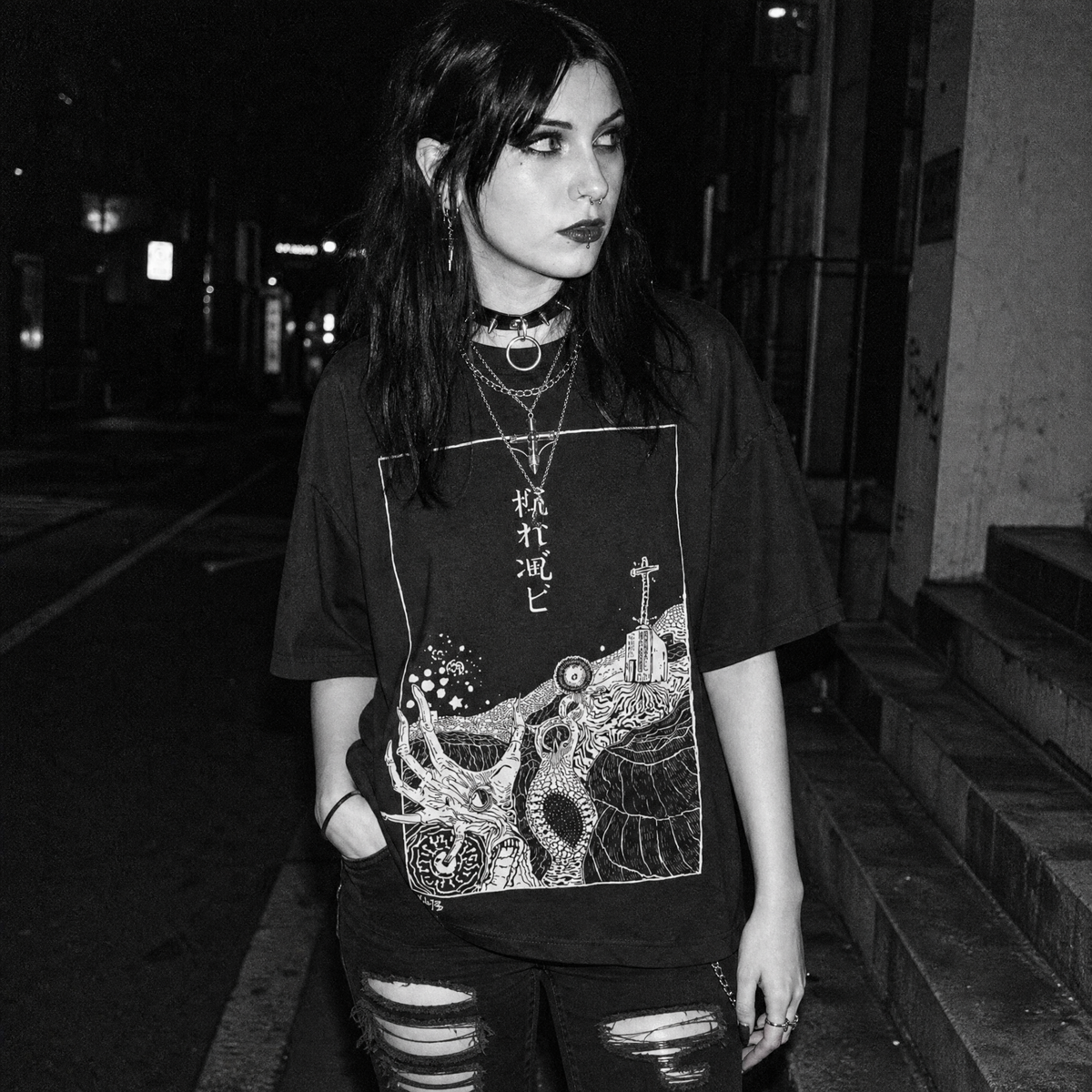 A woman standing on a city street at night wearing a black graphic t-shirt with a detailed white illustration of a deep sea horror scene and Japanese characters, paired with ripped jeans and layered necklaces.