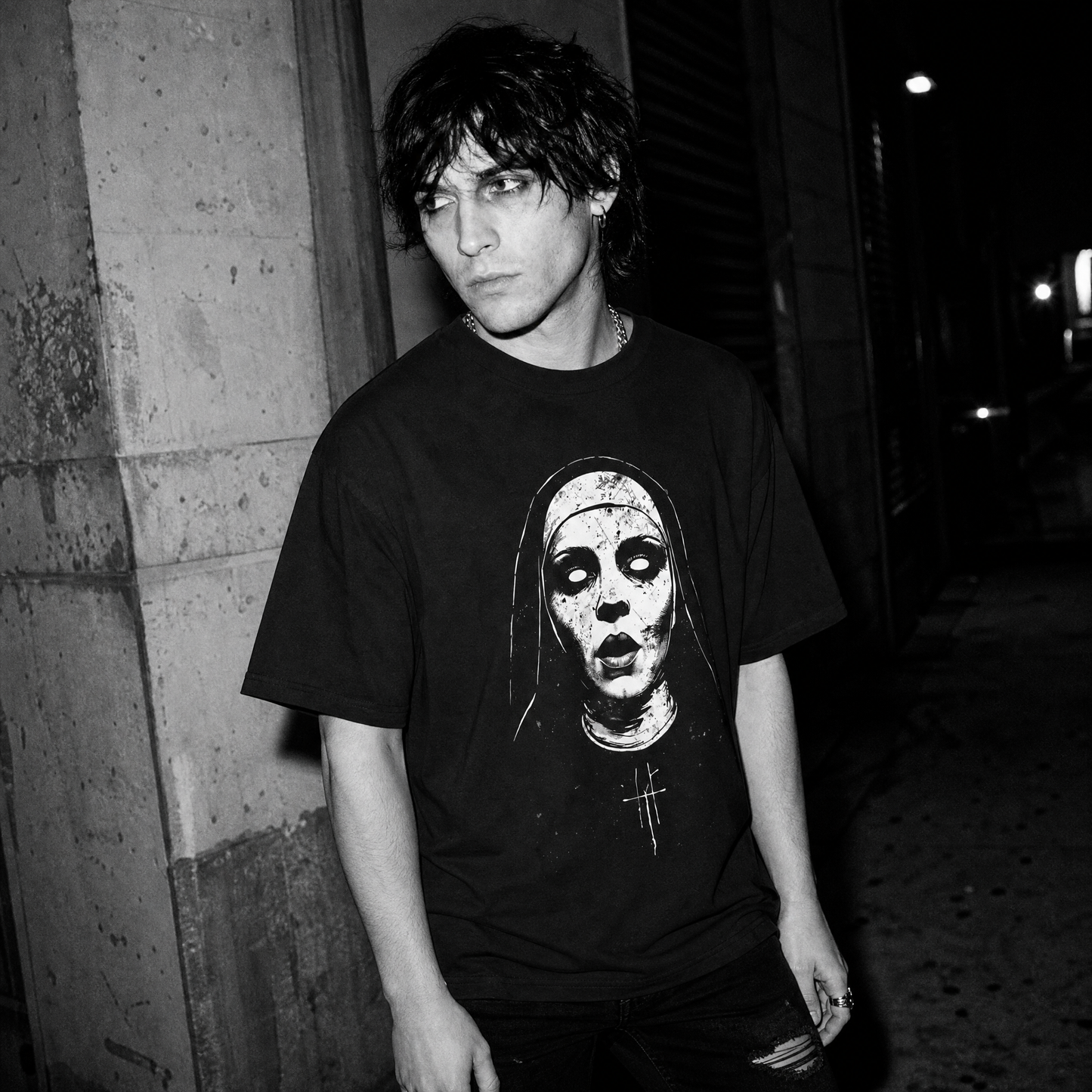 A person standing outdoors at night wearing a black graphic T-shirt with a white horror-style nun's face print on the front.