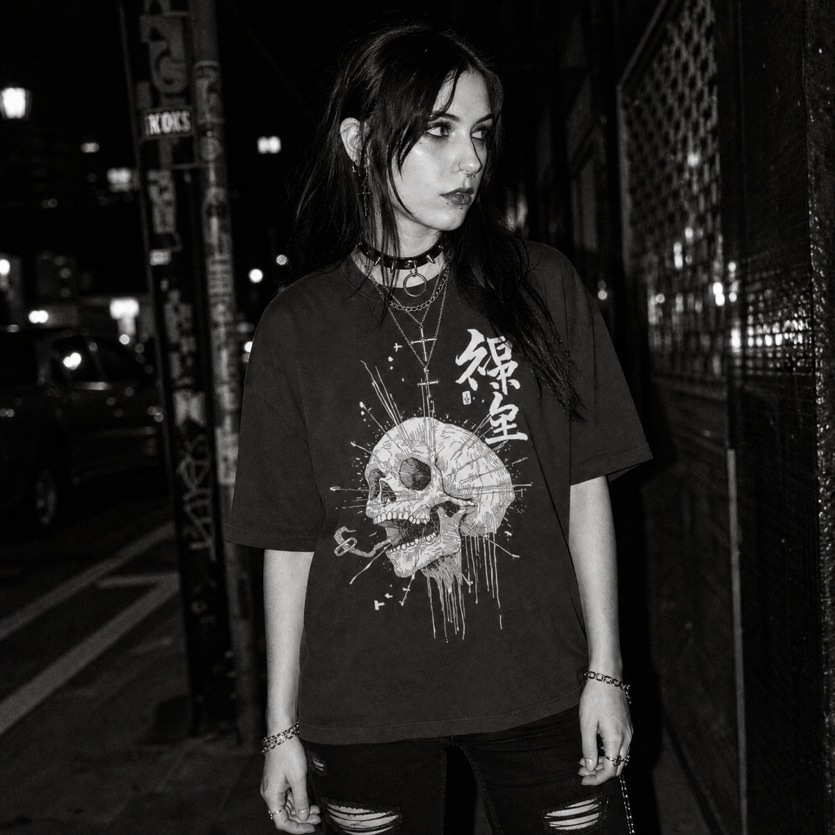 A woman standing outdoors at night wearing an oversized black graphic T-shirt with a large white skull print and Japanese characters on the front, paired with dark ripped jeans and layered necklaces.