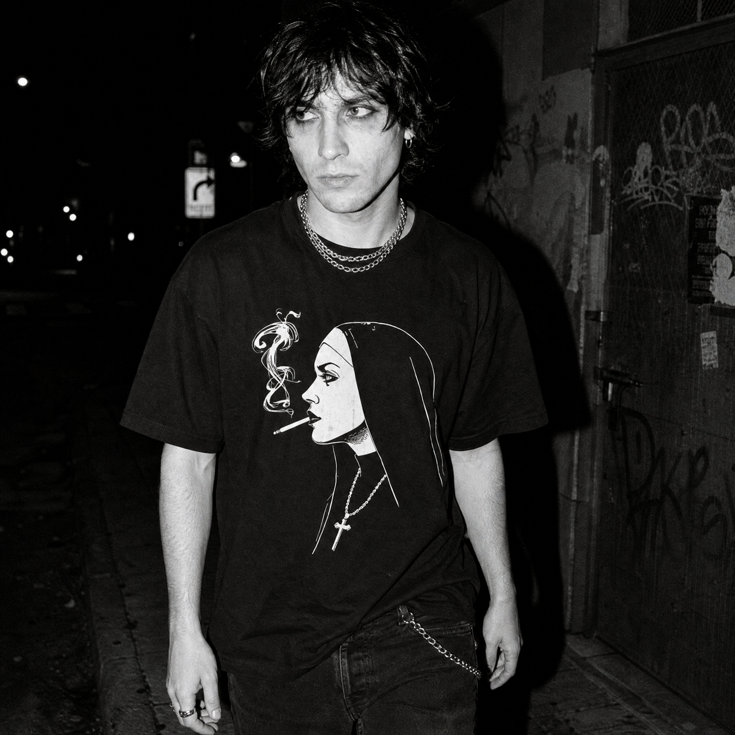 A black and white street-style photograph of a young man wearing a black graphic t-shirt with a white line art illustration of a smoking nun, standing in a dark outdoor alleyway.