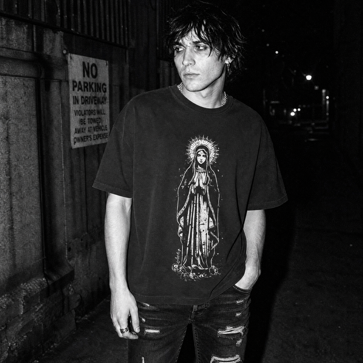 A man wearing a black graphic t-shirt featuring a detailed white line art illustration of the Virgin Mary, standing on a dark street at night.