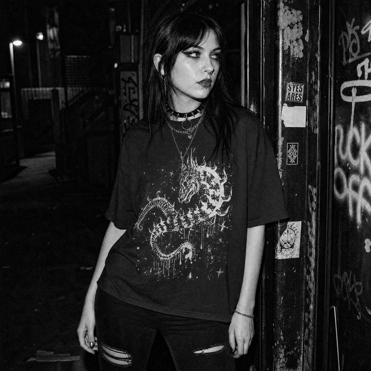 A woman stands in a dark alley at night wearing an oversized black graphic t-shirt with a dragon amidst dripping paint and splattered stars, paired with ripped black jeans and layered necklaces.