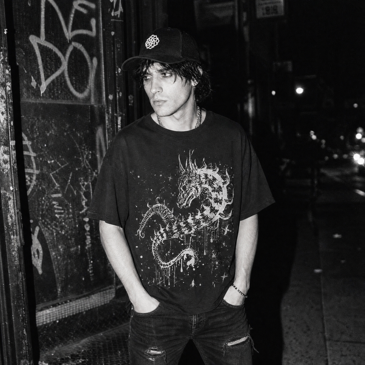 A man wearing a black t-shirt with a white distressed dragon amidst dripping paint and splattered stars, a black baseball cap, and ripped jeans, standing on a dark city street at night.