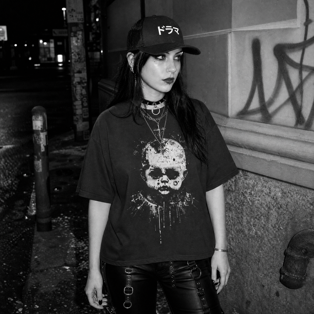 A woman standing outdoors at night wearing a black graphic T-shirt with a distressed creepy doll print, black leather pants, layered necklaces, and a black baseball cap with white Japanese text.