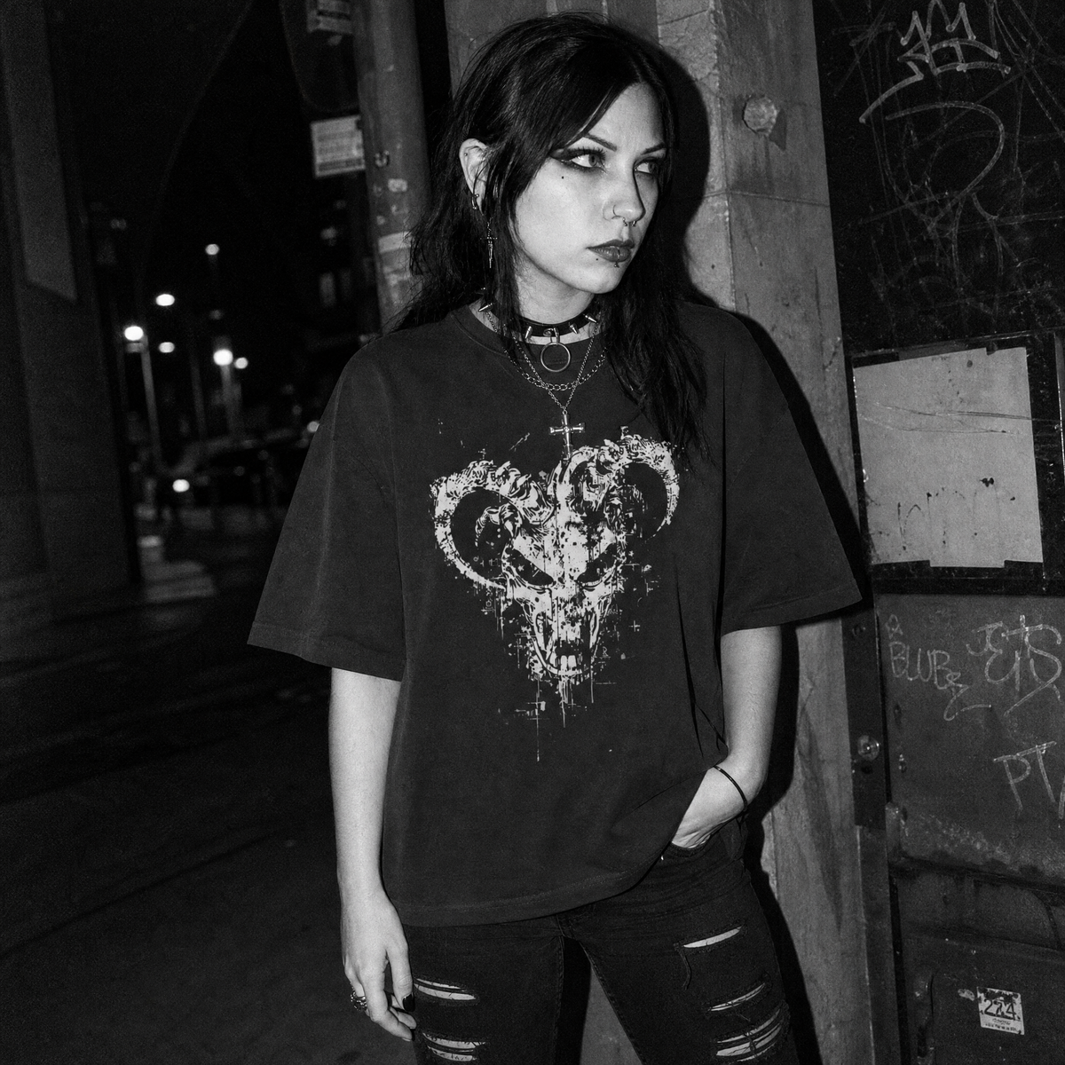 A woman wearing a black graphic t-shirt with a distressed white ram demon skull design, standing in a dark, gritty urban alleyway at night.