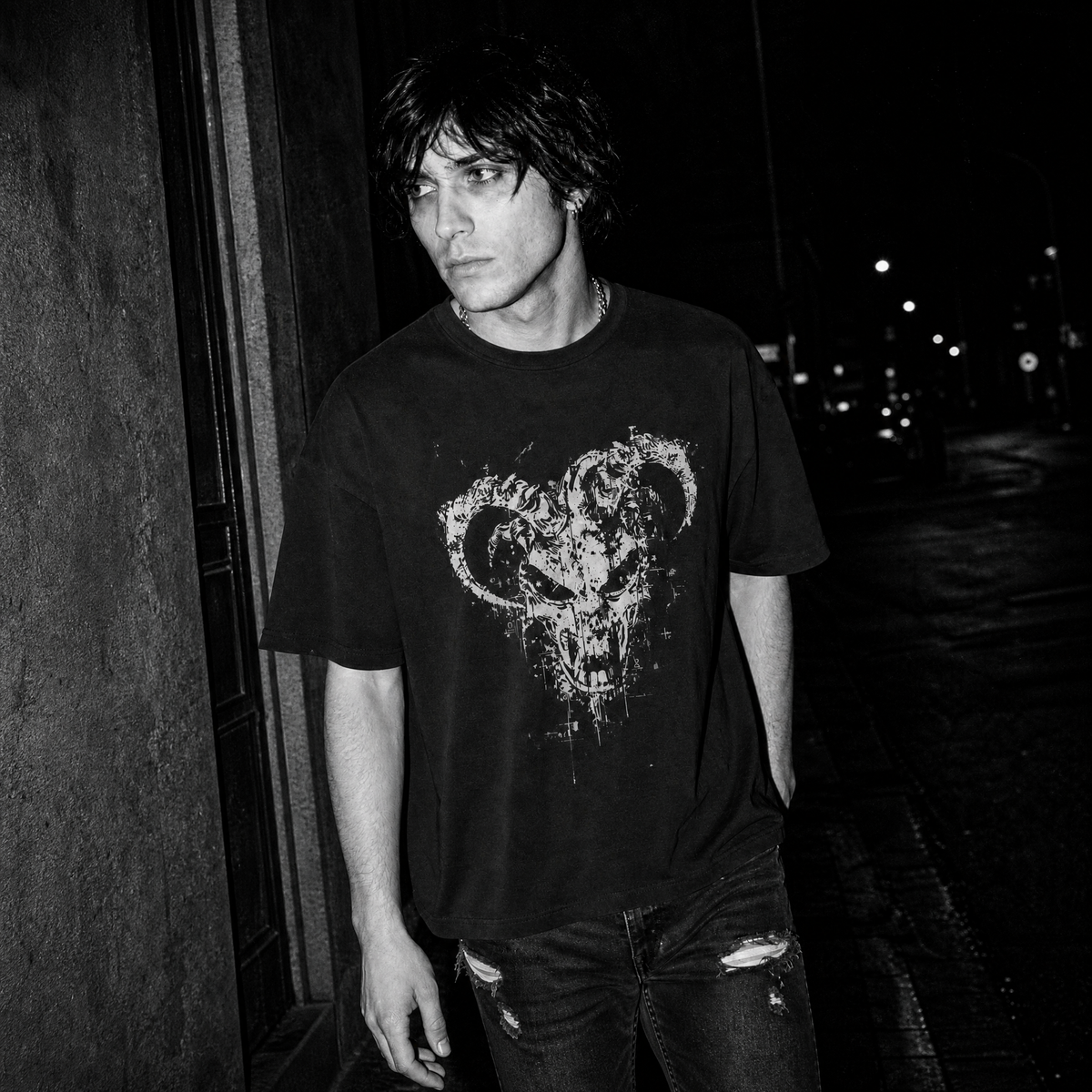 A man wearing a black graphic t-shirt with a distressed white ram demon skull design, standing in a dark outdoor night setting.