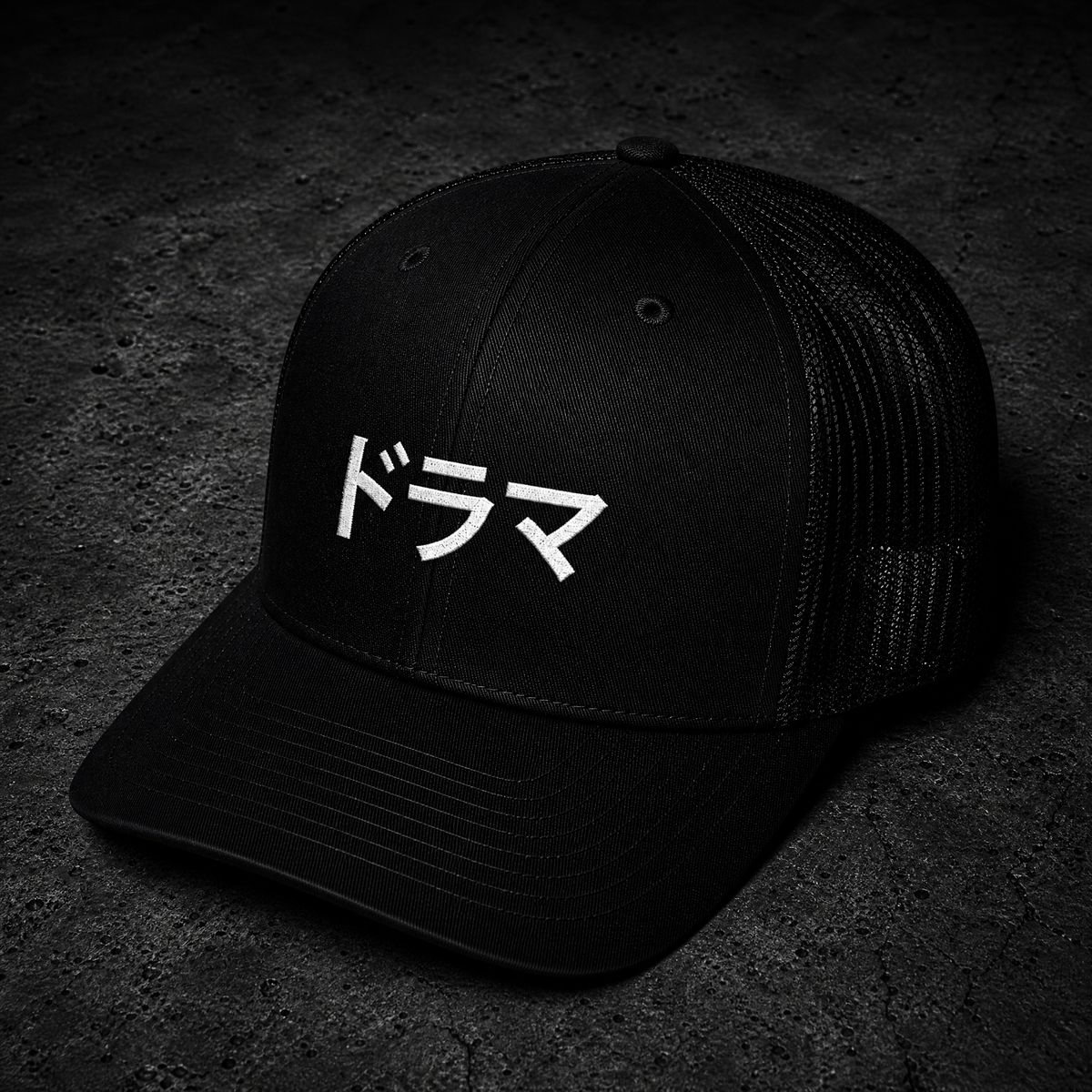 A black trucker-style cap with white Japanese text on the front that says DRAMA, photographed on a dark textured surface.