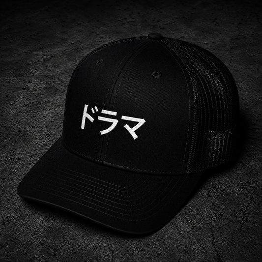 A black trucker-style cap with white Japanese text on the front that says DRAMA, photographed on a dark textured surface.