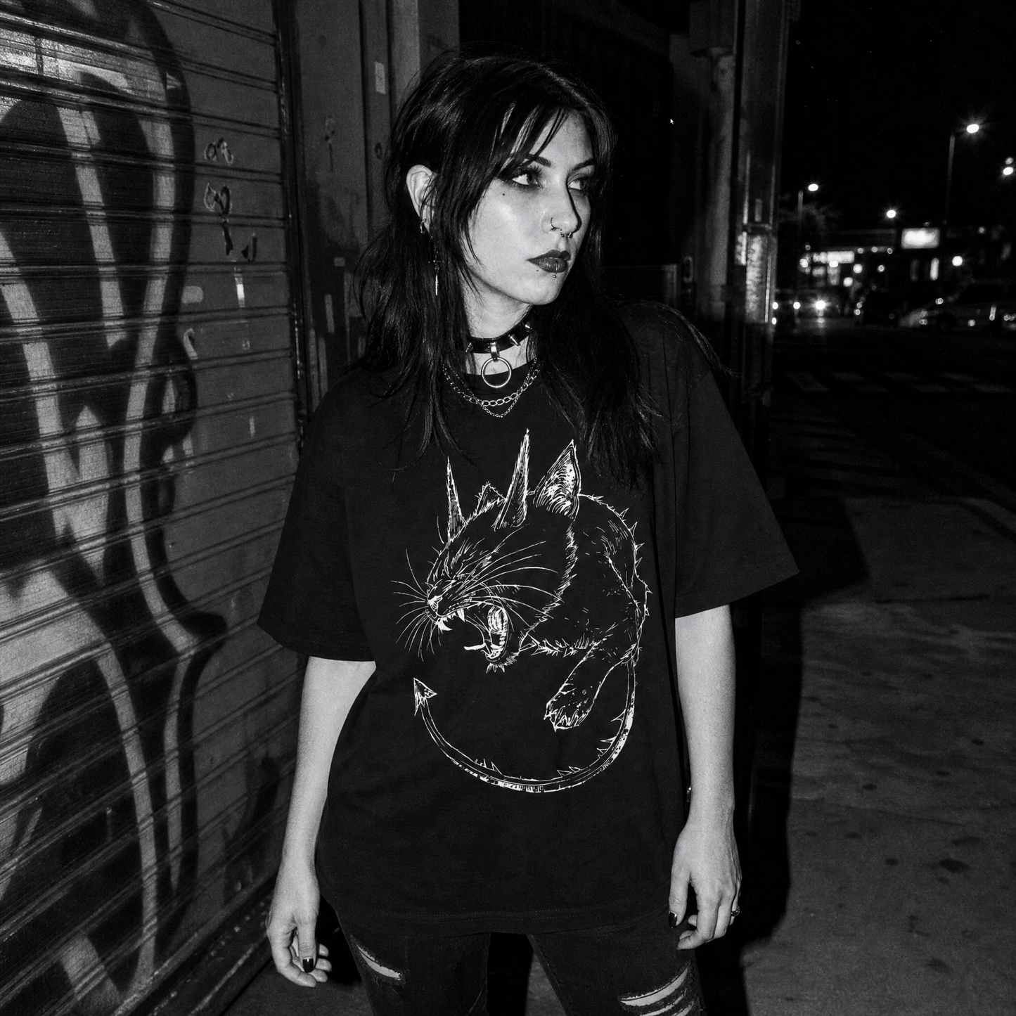 A woman wearing a black graphic t-shirt with a white demonic cat illustration, standing on a city street at night.