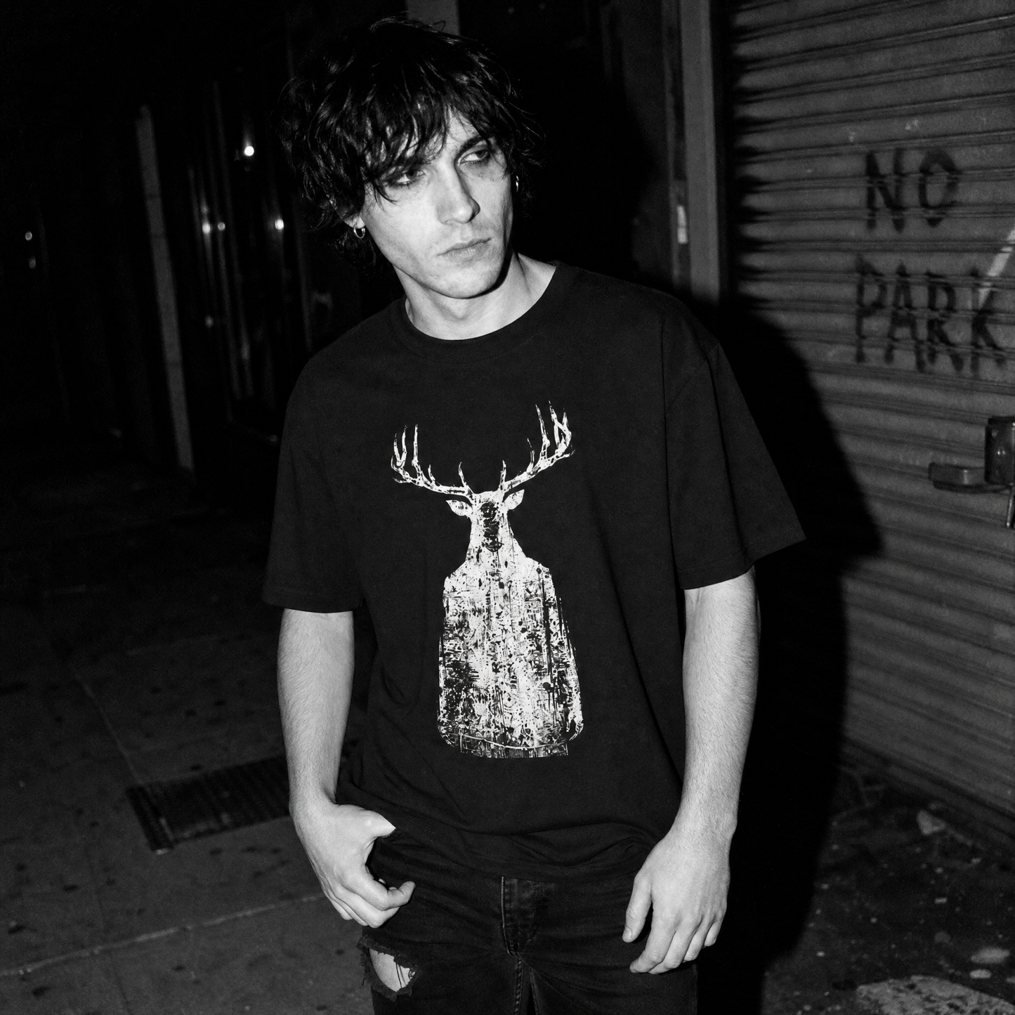 A black and white street-style photograph of a man wearing a black graphic t-shirt with a distressed print of a ghostly human with antlers, set against a dark urban alley background.