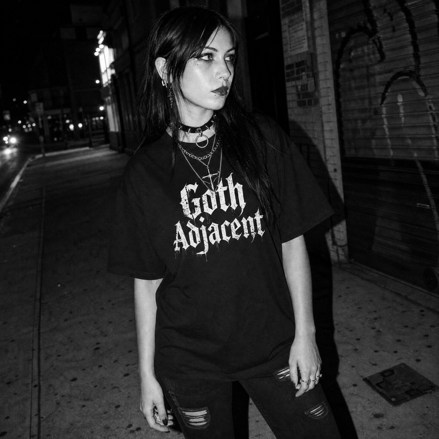 A woman posing on a city street at night wearing an oversized black t-shirt with 'Goth Adjacent' printed in a white gothic font, paired with distressed black jeans and layered necklaces.