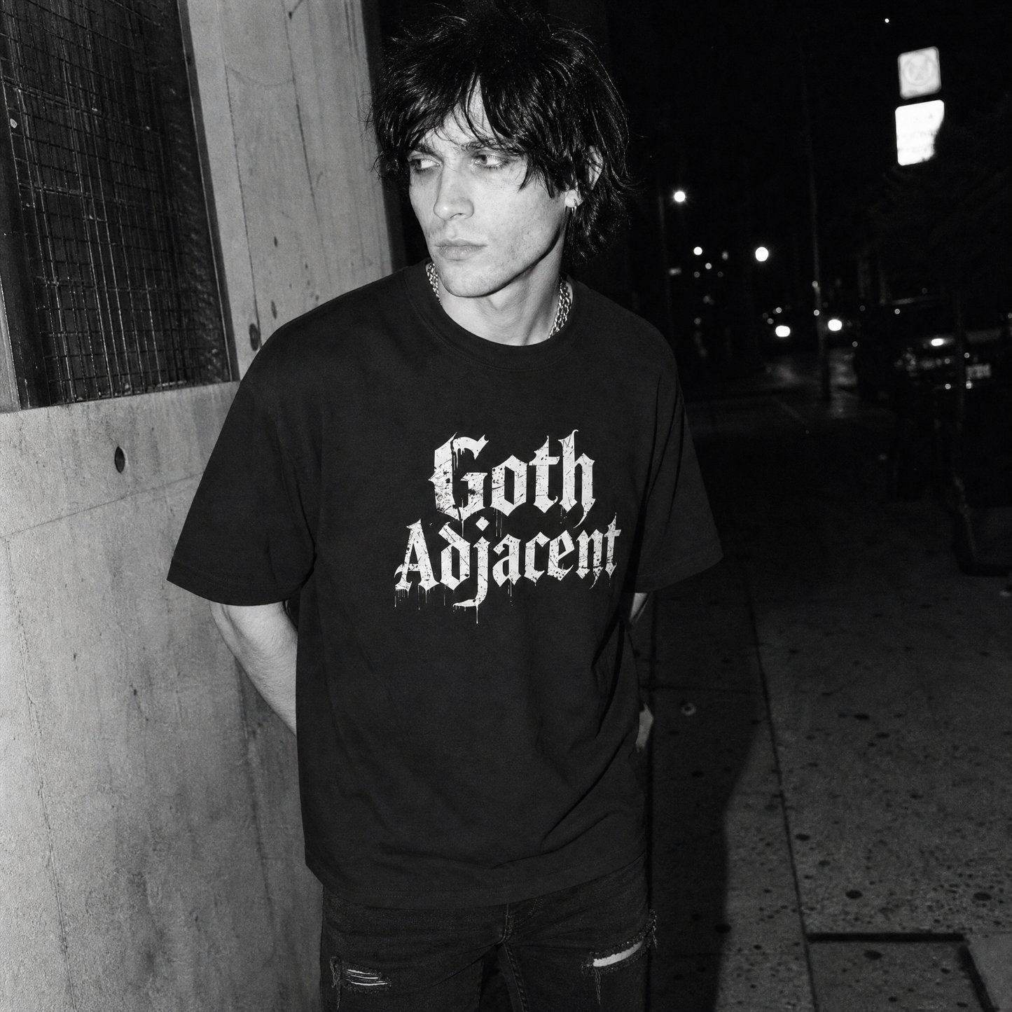 A man standing outdoors at night wearing a black graphic T-shirt with the words 'Goth Adjacent' in a white gothic font, paired with dark jeans and a chain necklace.
