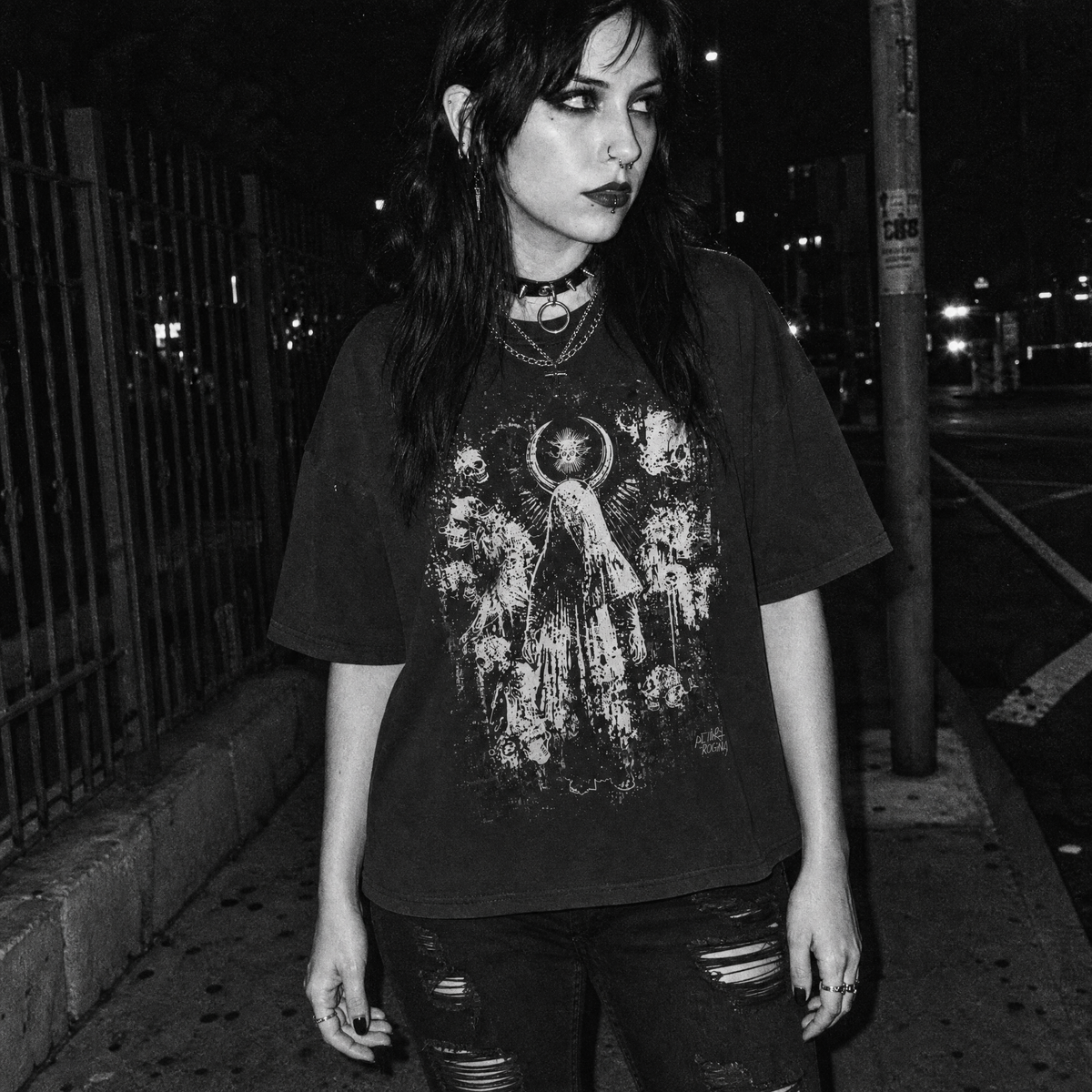 A woman stands on a city street at night wearing an oversized black graphic T-shirt with a distressed white print of a white angel, skull and crescent moon grunge artwork, paired with ripped dark jeans and layered necklaces.