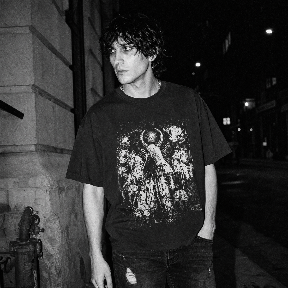 A man wearing a black graphic t-shirt with a distressed white print of a  distressed white angel, skull and crescent moon grunge artwork, standing on a dark city street at night.