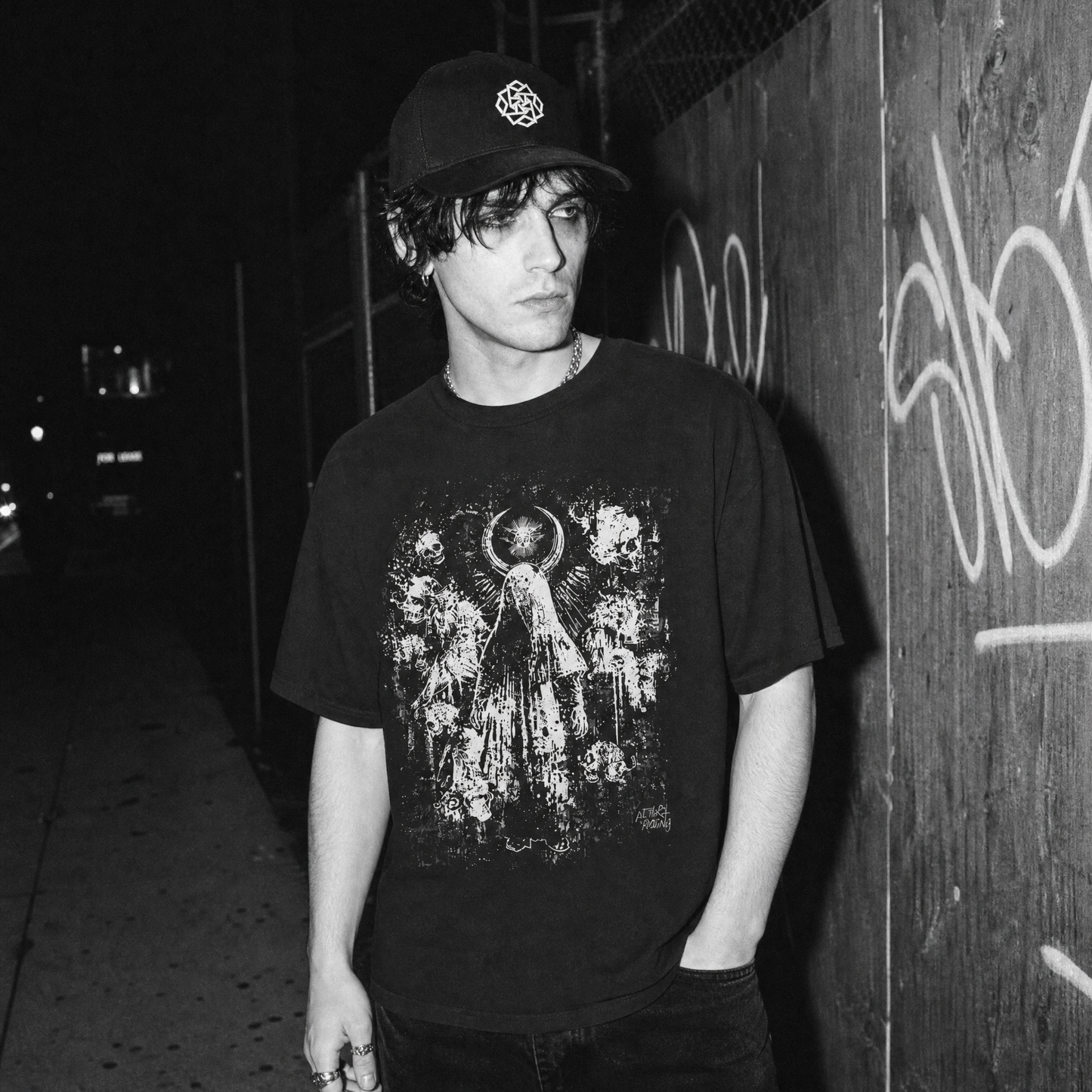 A man wearing a black graphic t-shirt with a distressed white angel print and a baseball cap stands against a graffiti-covered wooden wall at night.