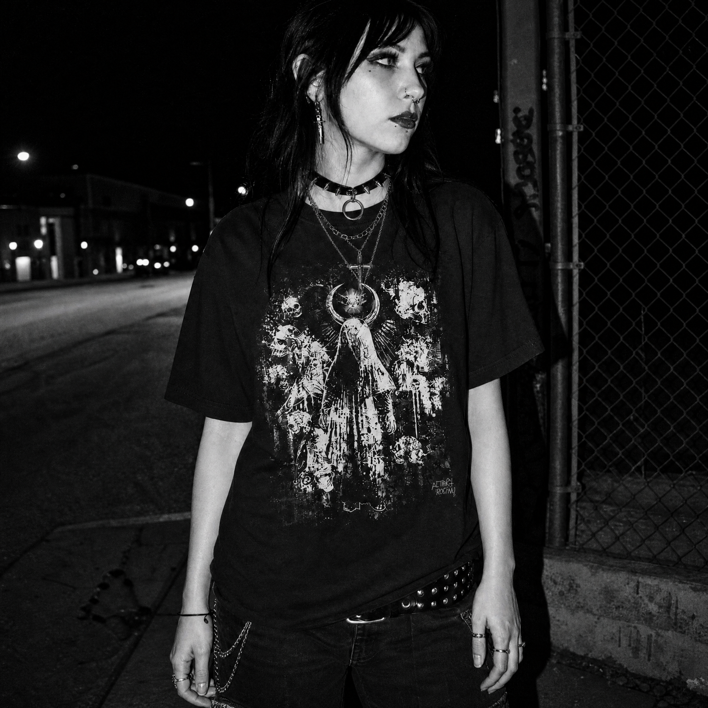 A female standing outdoors at night wearing a black graphic T-shirt with a distressed white angel print, layered necklaces, and dark jeans.