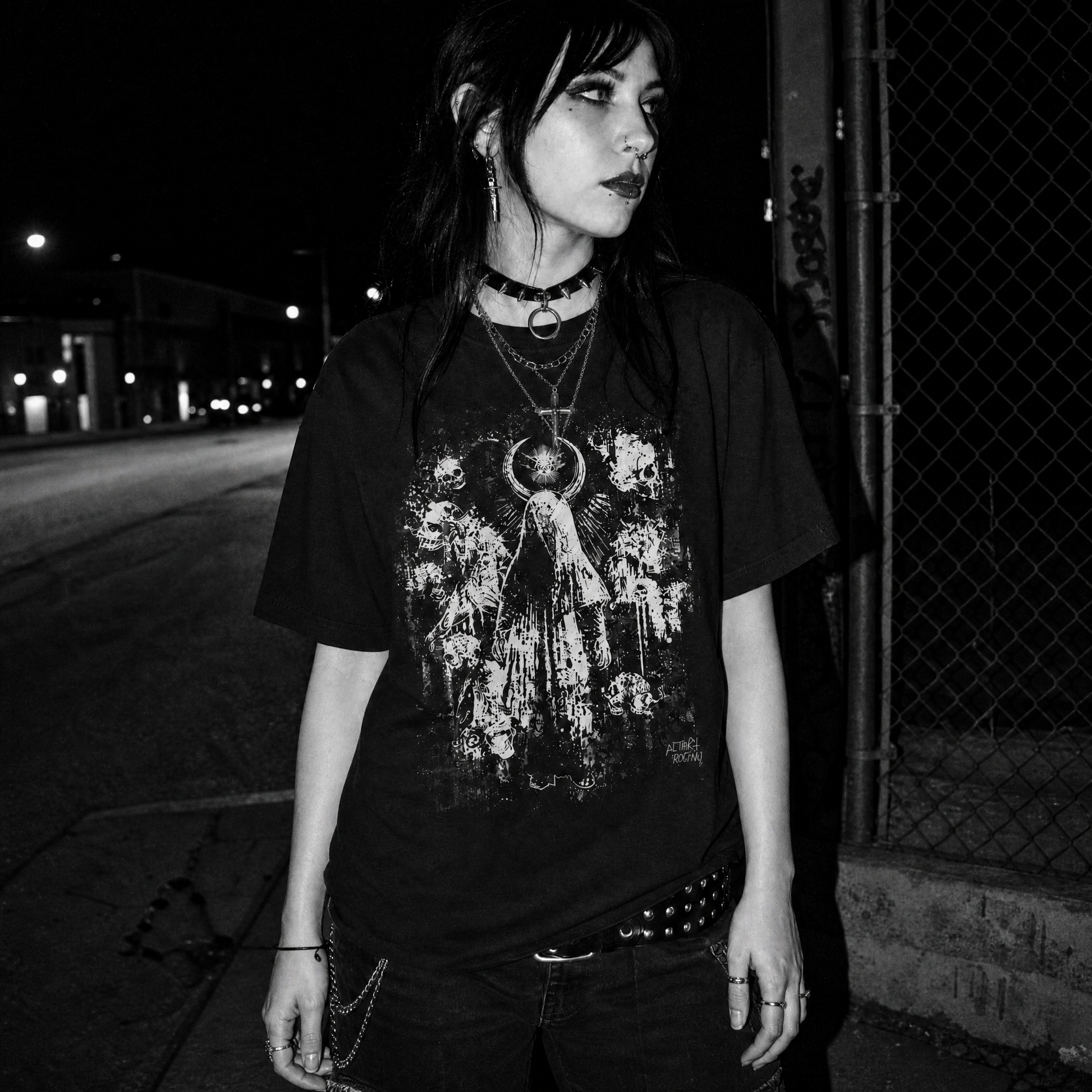 A female standing outdoors at night wearing a black graphic T-shirt with a distressed white angel print, layered necklaces, and dark jeans.