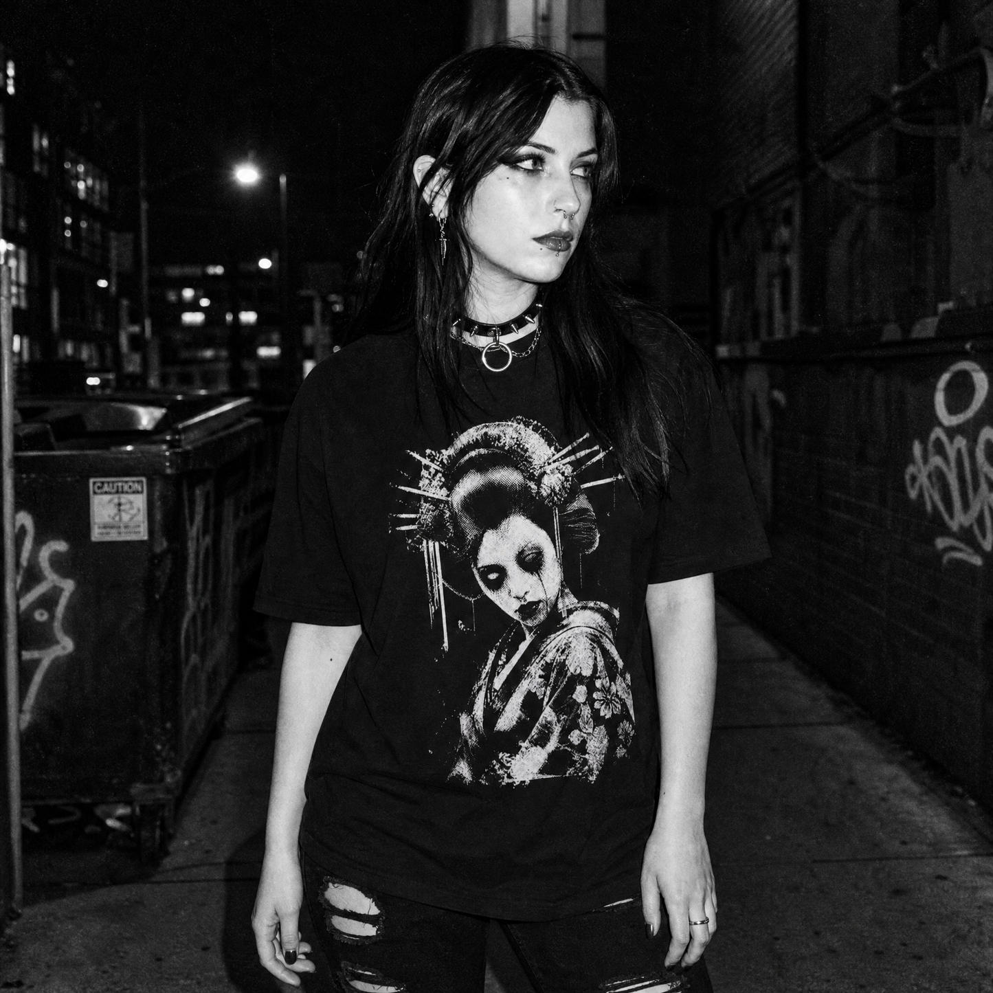A woman stands on a dark urban sidewalk at night wearing a black graphic T-shirt with high-contrast white graphic print of a geisha with traditional hairpins, makeup and dead eyes, along with ripped jeans and layered necklaces.