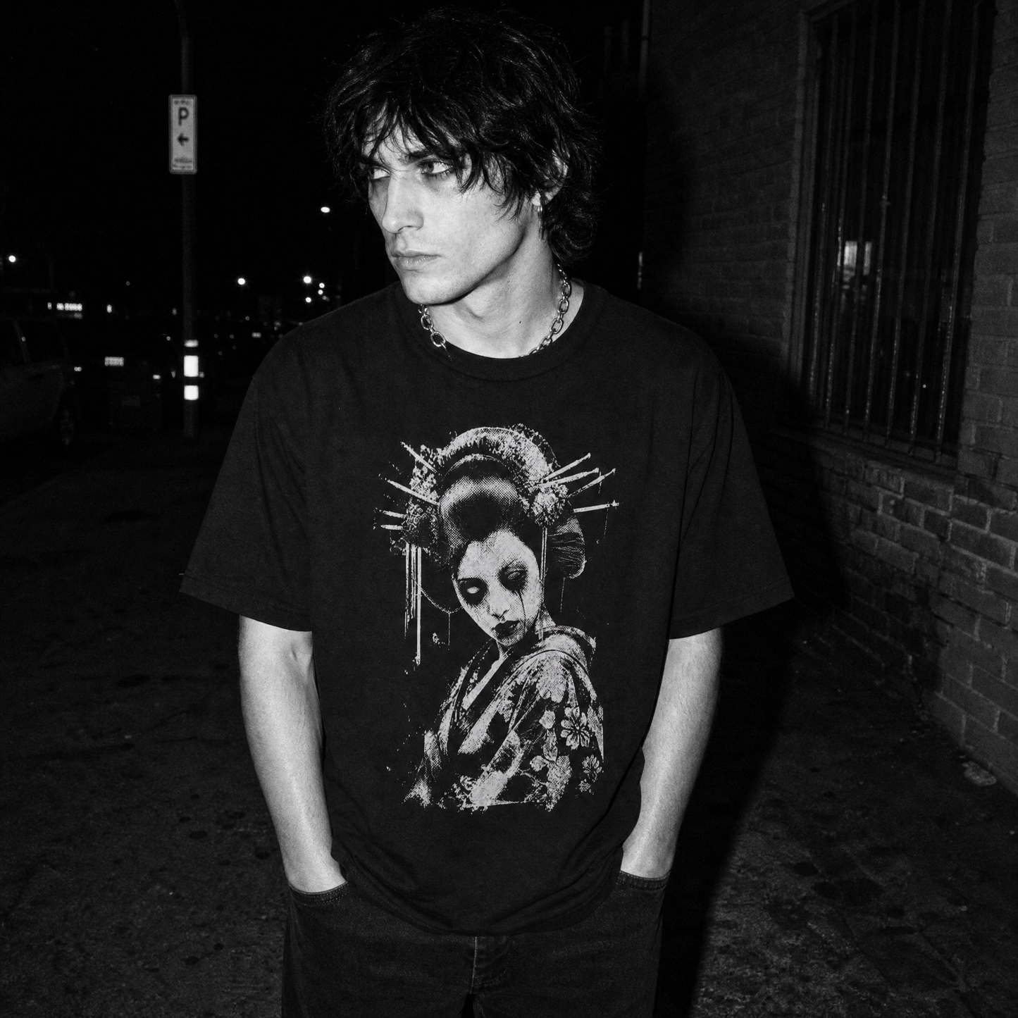 A man wearing a black graphic t-shirt with a high-contrast white graphic print of a geisha with traditional hairpins, makeup and dead eyes, standing outdoors at night against a brick wall.