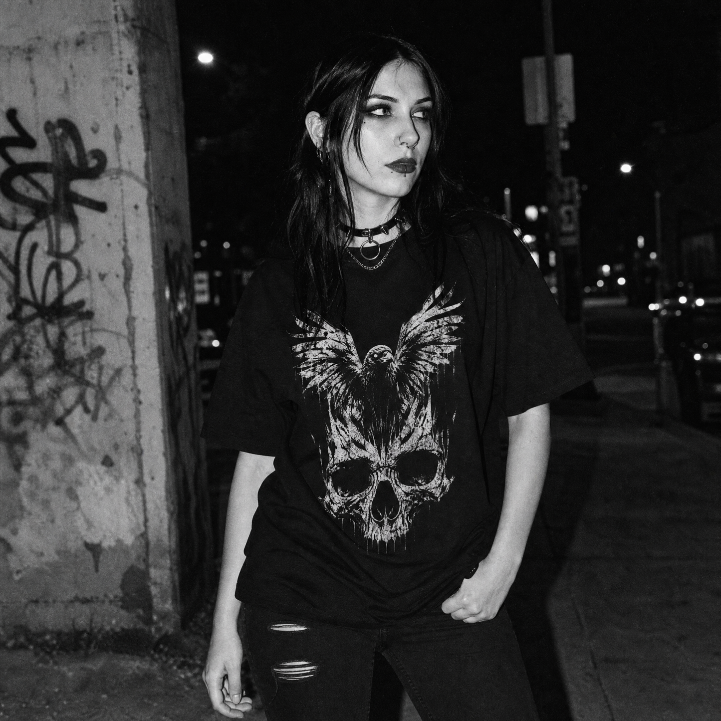 A woman wearing a black graphic t-shirt with a large white skull and raven design, standing on a city sidewalk at night.