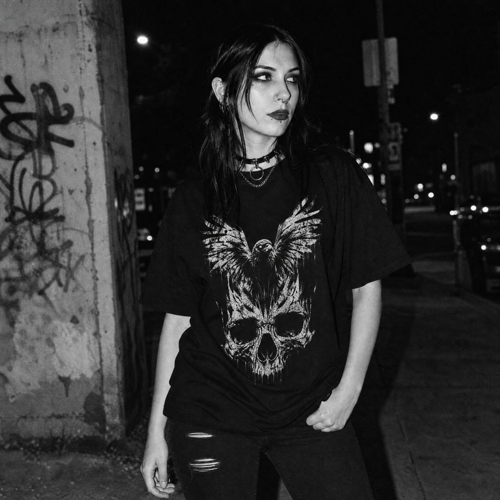 A woman wearing a black graphic t-shirt with a large white skull and raven design, standing on a city sidewalk at night.