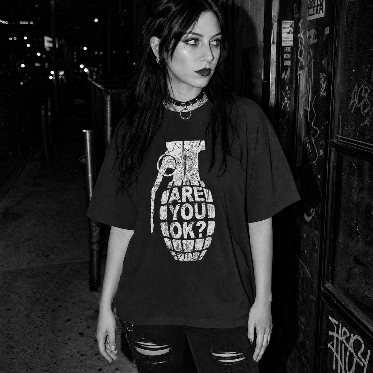 A woman wearing a black oversized graphic t-shirt with a white grenade print that reads 'ARE YOU OK?' stands on a dark street at night.
