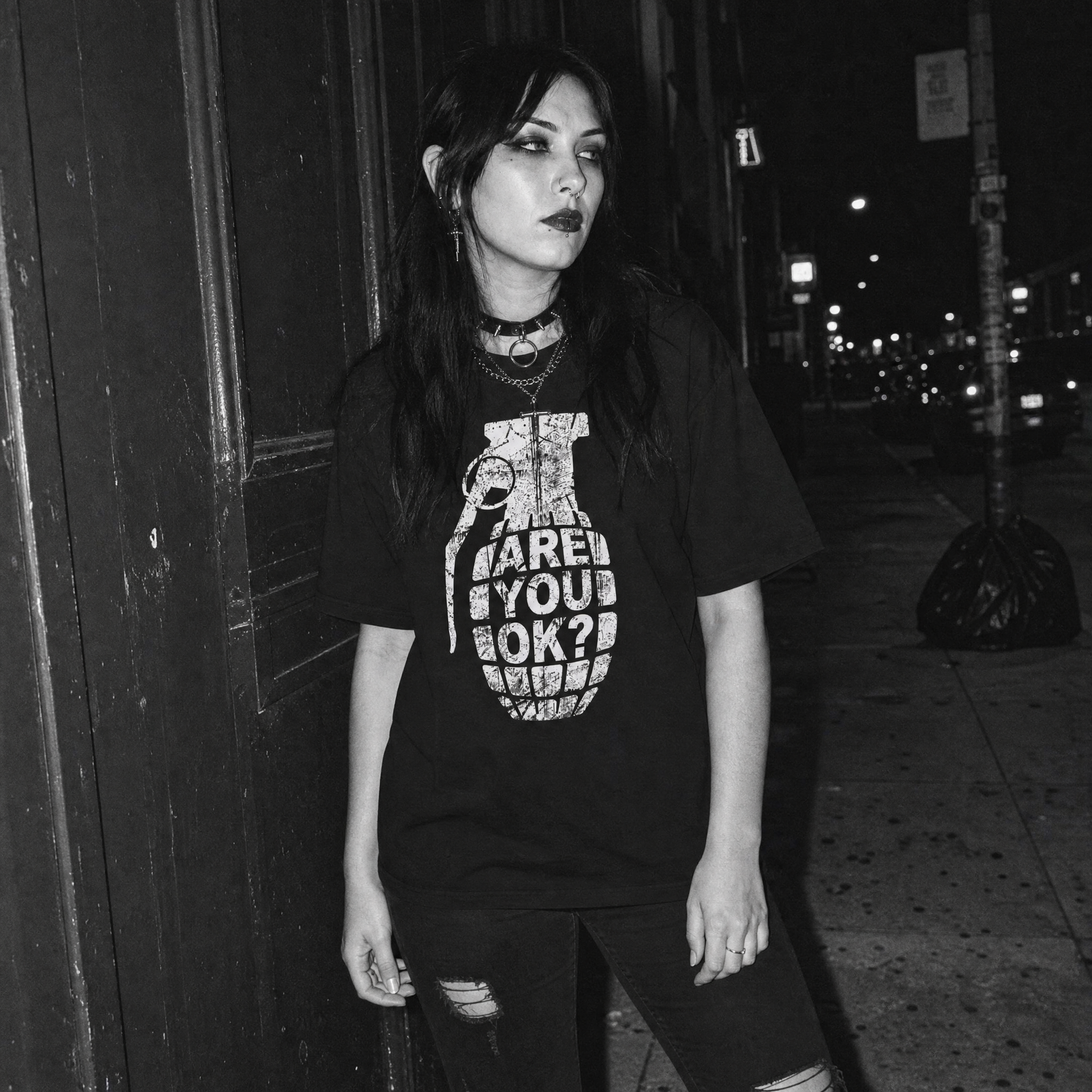 A woman wearing a black graphic t-shirt with a white grenade design and the text 'ARE YOU OK?' stands on a city sidewalk at night.