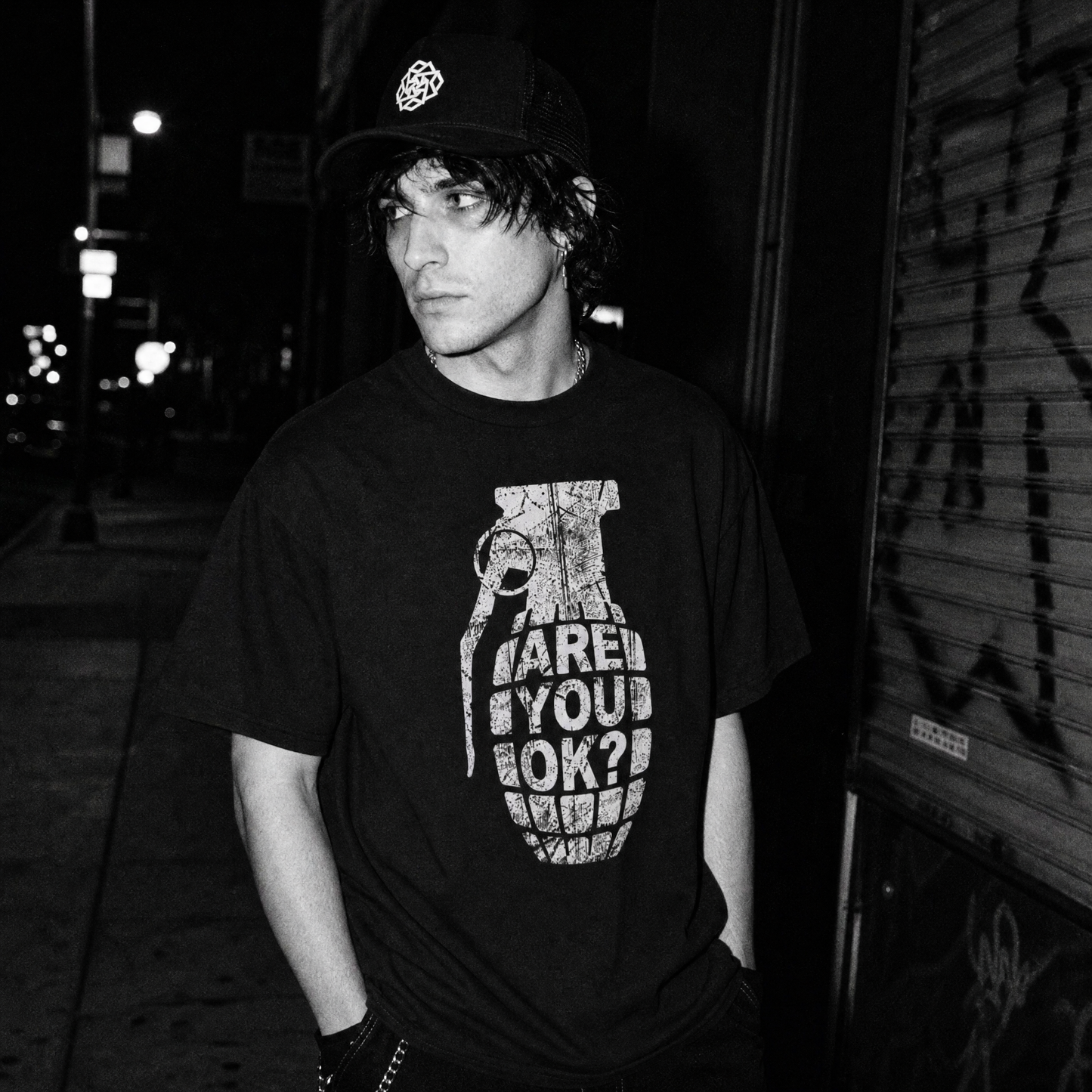 A man wearing a black graphic t-shirt with a white grenade print and the text 'ARE YOU OK?', standing in a dark street corner at night.