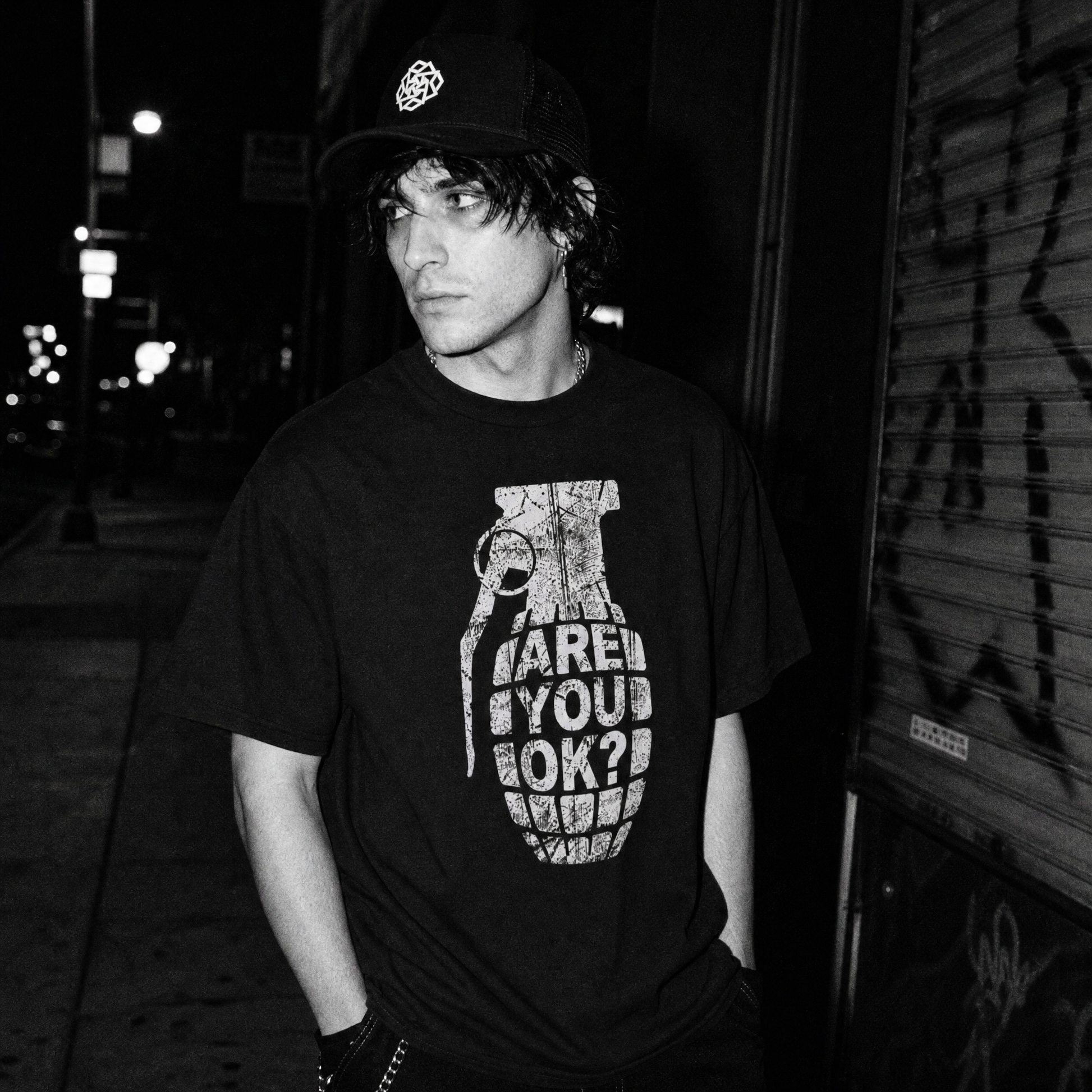 A man wearing a black graphic t-shirt with a white grenade print and the text 'ARE YOU OK?', standing in a dark street corner at night.
