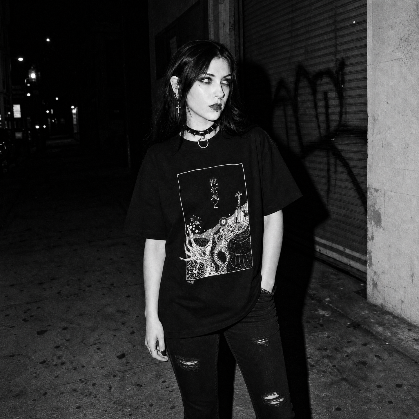 A woman stands at night wearing a black graphic t-shirt with a stylized deep sea horror image and white Japanese-style Kanji print and distressed black jeans, photographed in a dark urban setting.