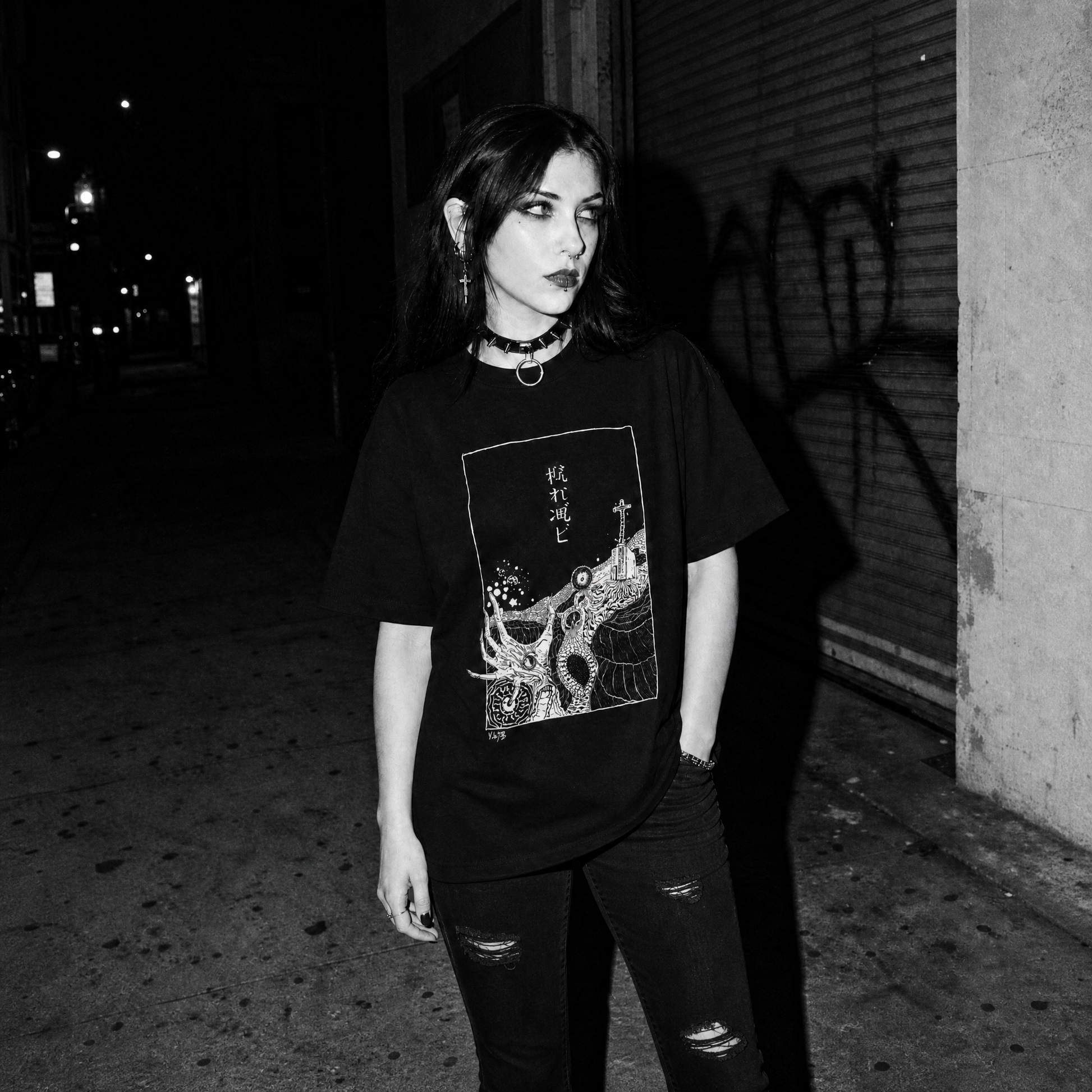 A woman stands at night wearing a black graphic t-shirt with a stylized deep sea horror image and white Japanese-style Kanji print and distressed black jeans, photographed in a dark urban setting.