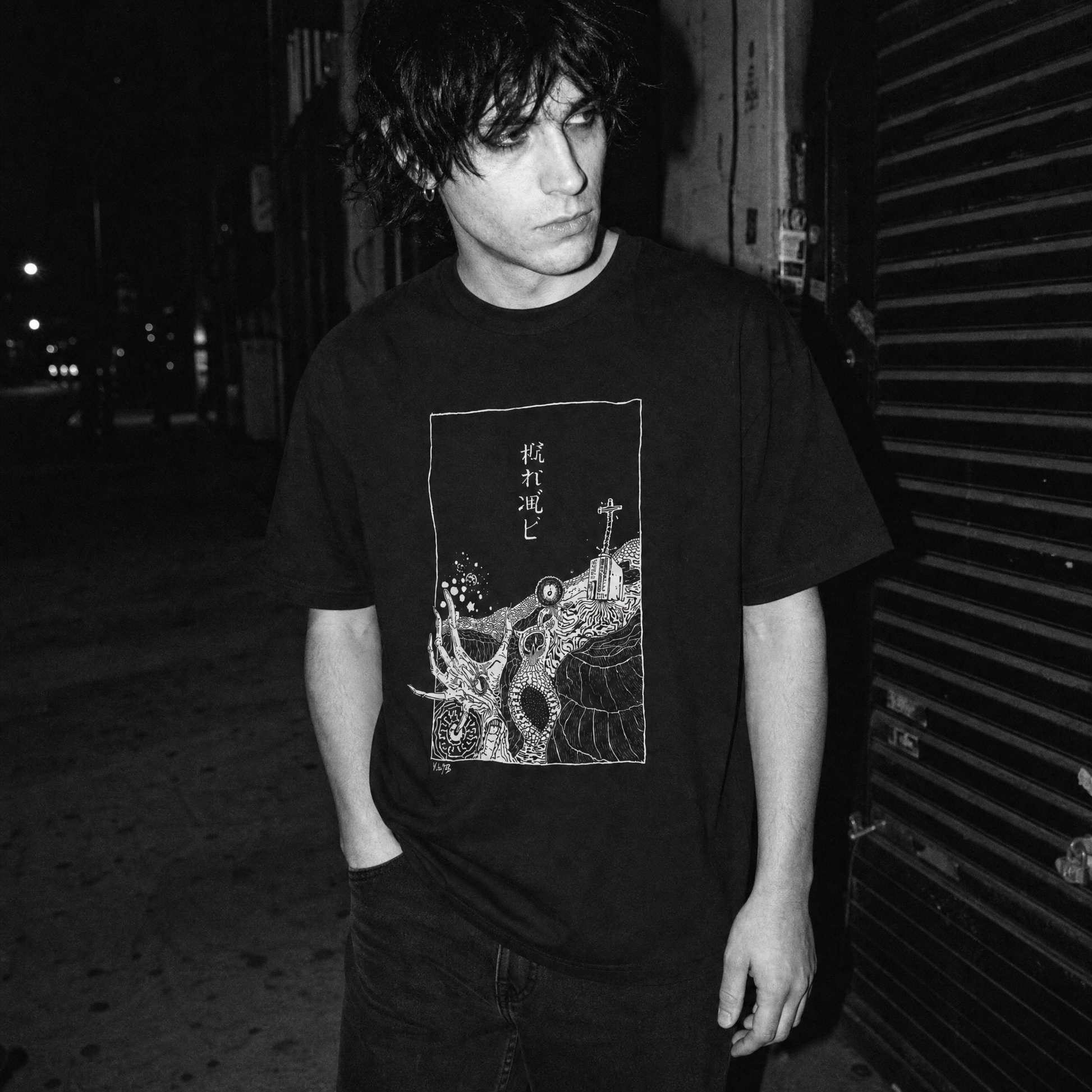 A man wearing a black graphic t-shirt with a stylized deep sea horror illustration and Japanese Kanji text, standing in a dark outdoor alley at night.