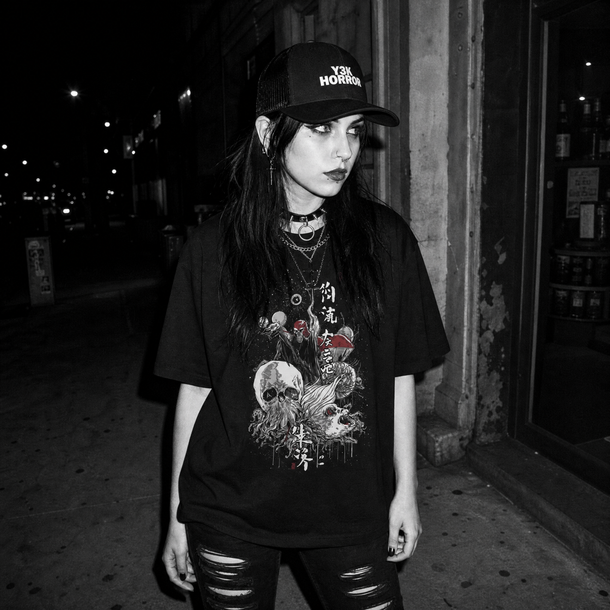 A woman wearing a black graphic t-shirt with a cosmic war and Japanese kanji print, a black baseball cap with 'Y3K HORROR' text, and ripped black jeans, standing on a dark city street at night.