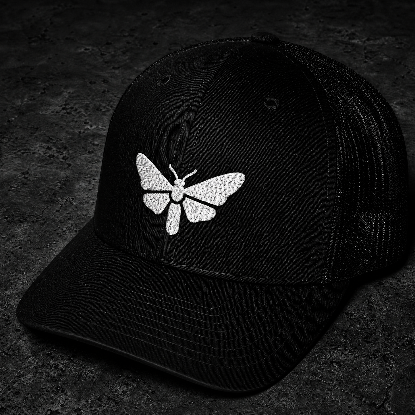 A black trucker-style cap with a white embroidered moth motif on the front, photographed on a dark textured surface.