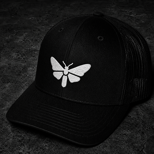 A black trucker-style cap with a white embroidered moth motif on the front, photographed on a dark textured surface.