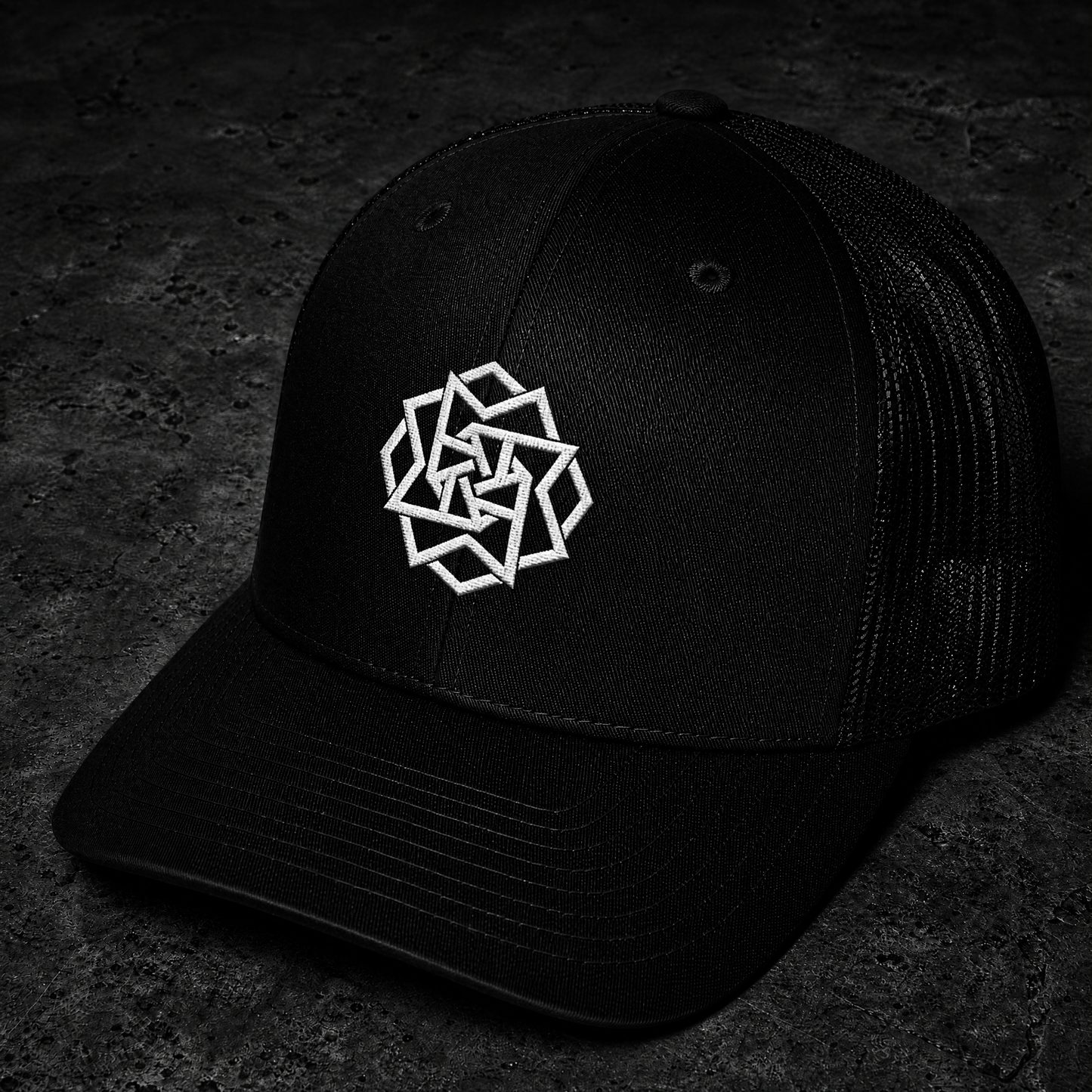 A black trucker-style baseball cap with a white geometric logo embroidered on the front, photographed on a dark textured surface.