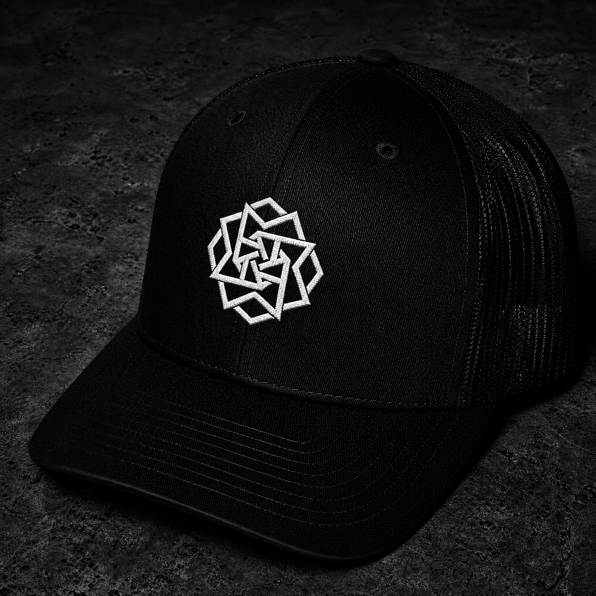 A black trucker-style baseball cap with a white geometric logo embroidered on the front, photographed on a dark textured surface.