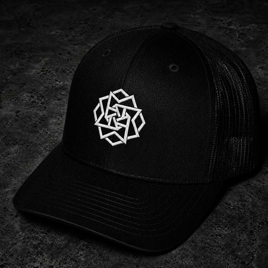 A black trucker-style baseball cap with a white geometric logo embroidered on the front, photographed on a dark textured surface.