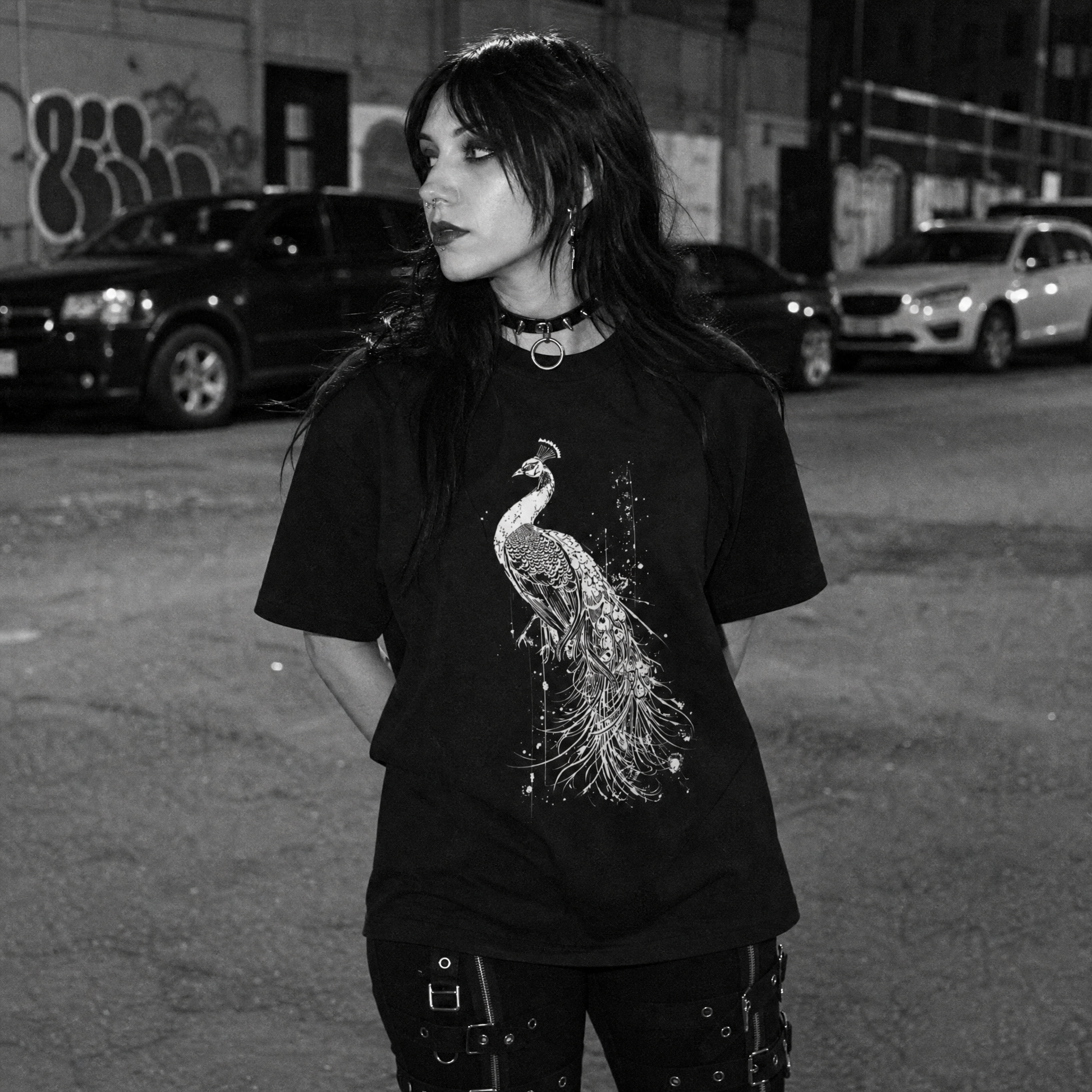 A woman wearing a black graphic t-shirt with a detailed white peacock print, standing in an urban street setting at night.