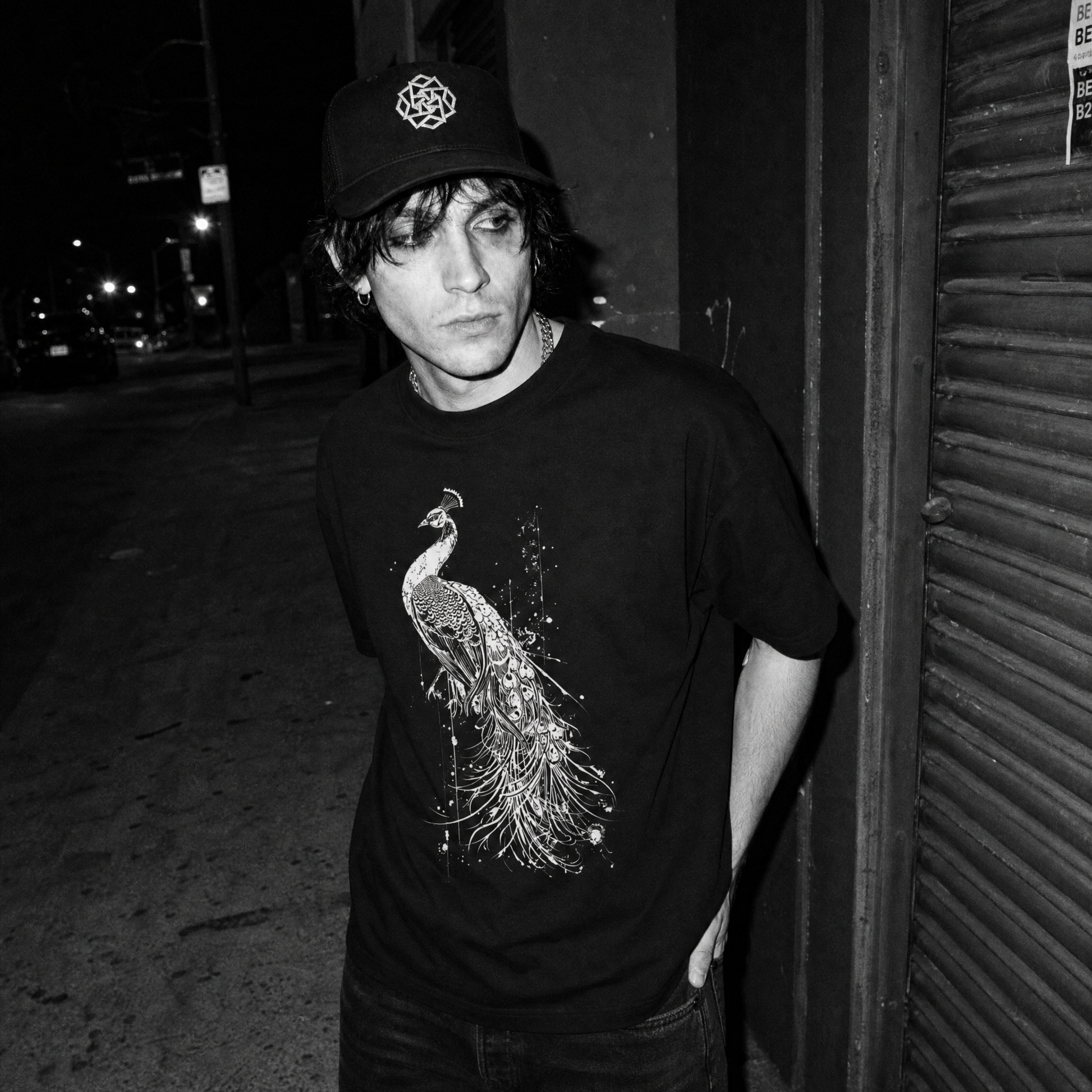 A black and white street-style photograph of a young man wearing a black t-shirt with a detailed peacock graphic, a black baseball cap, and dark jeans, standing next to a closed metal shutter at night.
