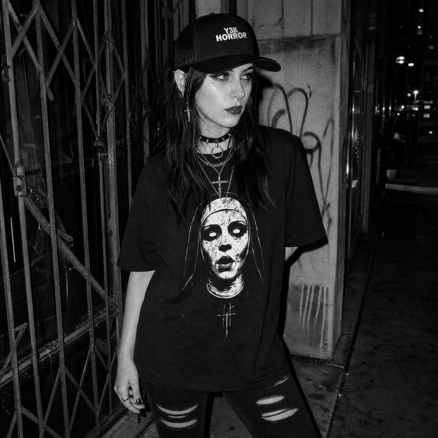 A woman wearing a black graphic t-shirt with a horror nun print, a black trucker cap with 'Y3K HORROR' text, and distressed jeans, standing on a dark city street at night.