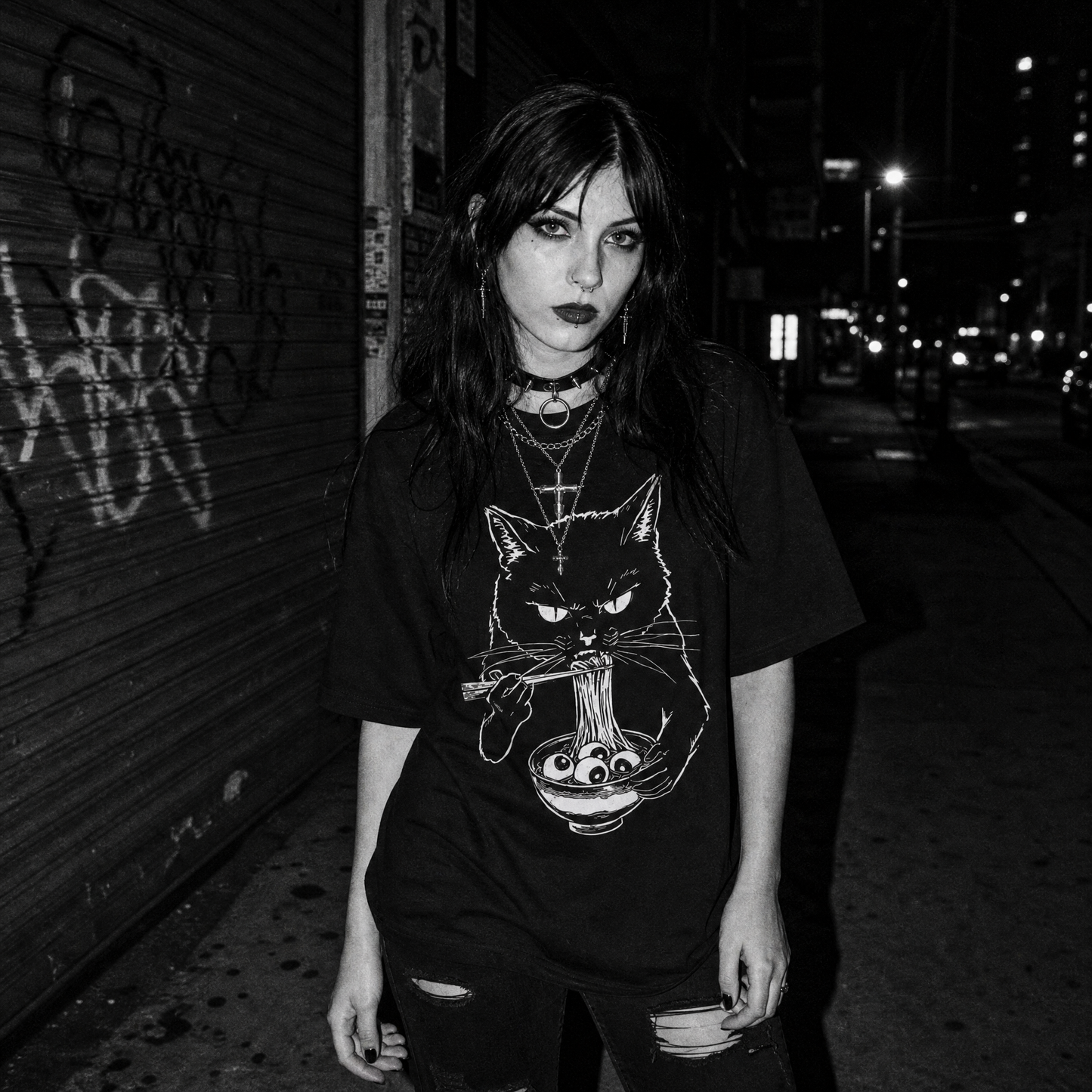 A woman stands on a dark city street at night wearing a black graphic T-shirt with a white cat illustration holding a bowl and eating noodles, paired with dark ripped jeans and layered necklaces.