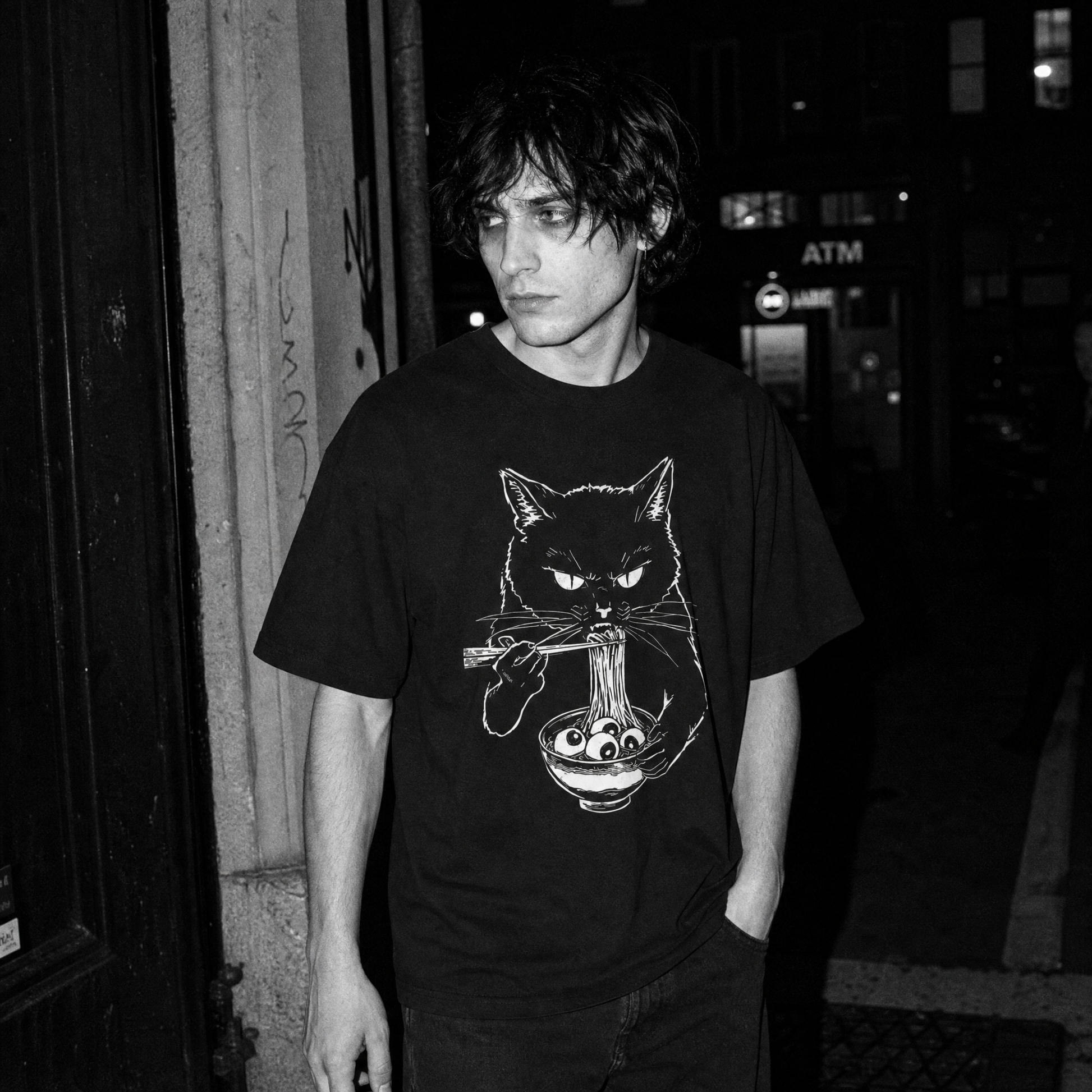 A man wearing a black graphic t-shirt with a white line art illustration of a cat eating ramen, standing in a dark, urban street setting.