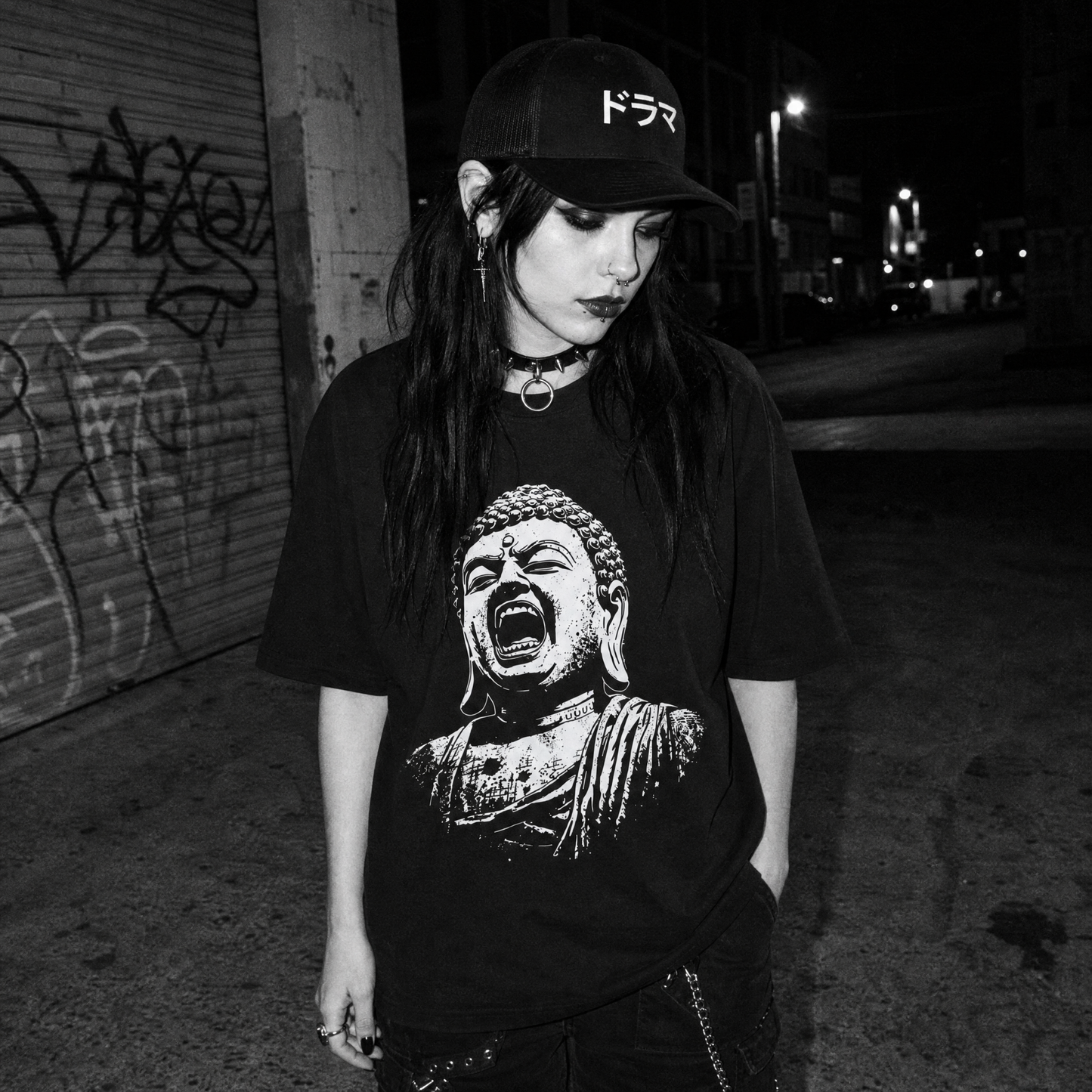 A woman stands on a city street at night wearing a black baseball cap with white Japanese text, a black graphic T-shirt featuring a screaming Buddha print, layered necklaces, and dark pants. The background shows a graffiti-covered shutter and street signs.