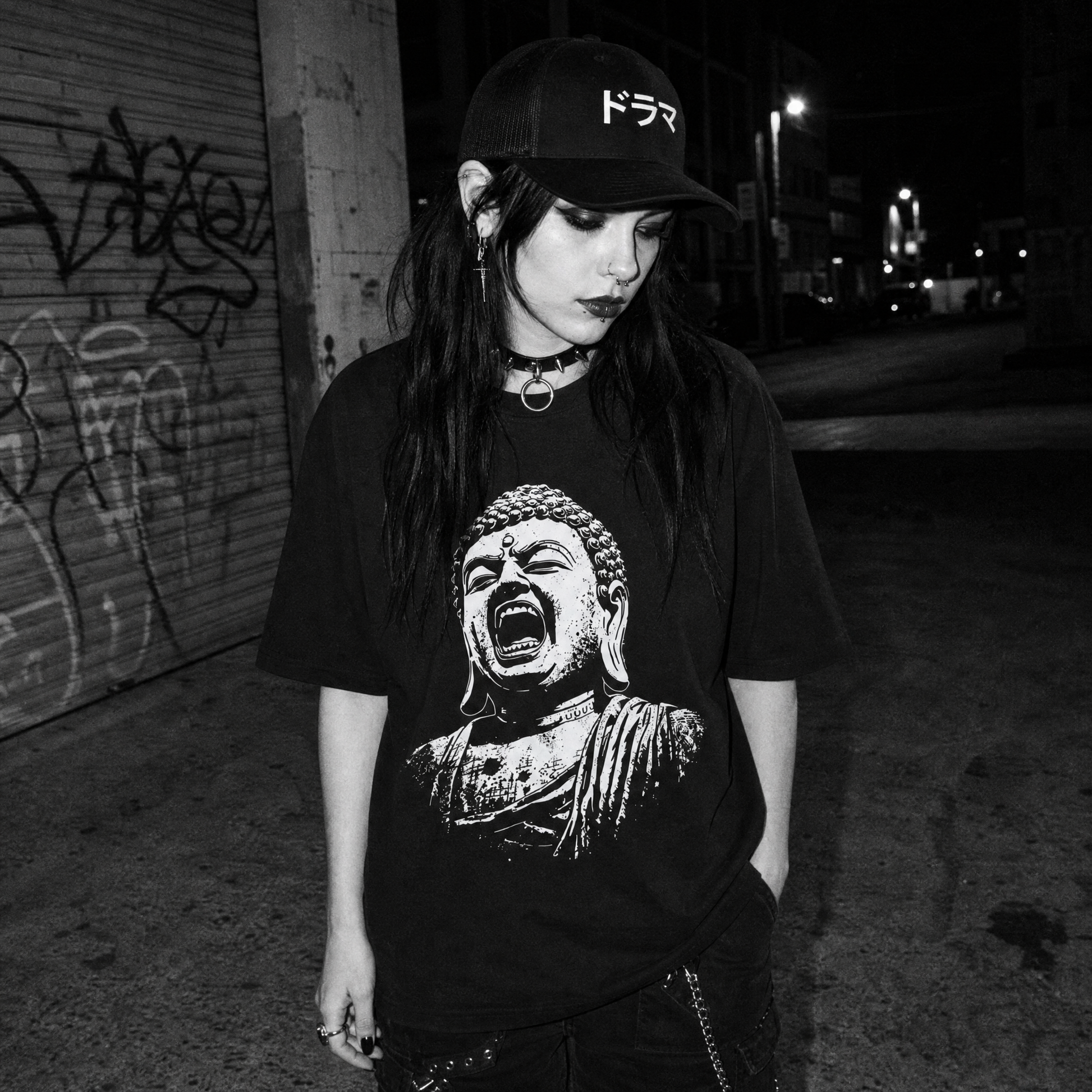A woman stands on a city street at night wearing a black baseball cap with white Japanese text, a black graphic T-shirt featuring a screaming Buddha print, layered necklaces, and dark pants. The background shows a graffiti-covered shutter and street signs.