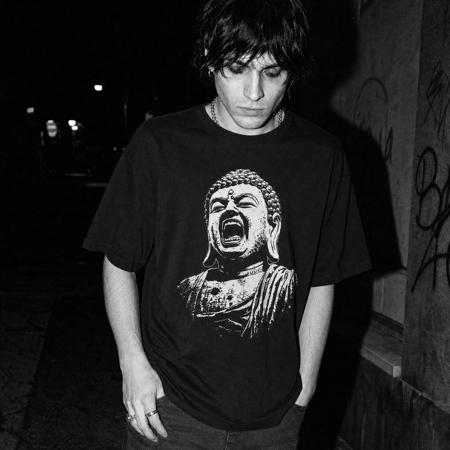 A male wearing a black short-sleeve graphic T-shirt with a large white screaming Buddha bust print on the front, photographed against a dark brick and metal gate background.